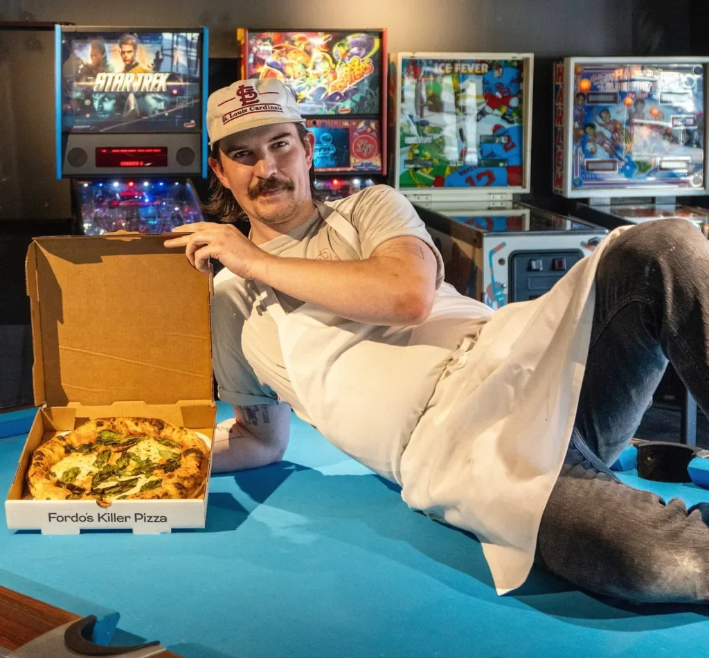 Chef on pool table with open pizza box and pinball machines.