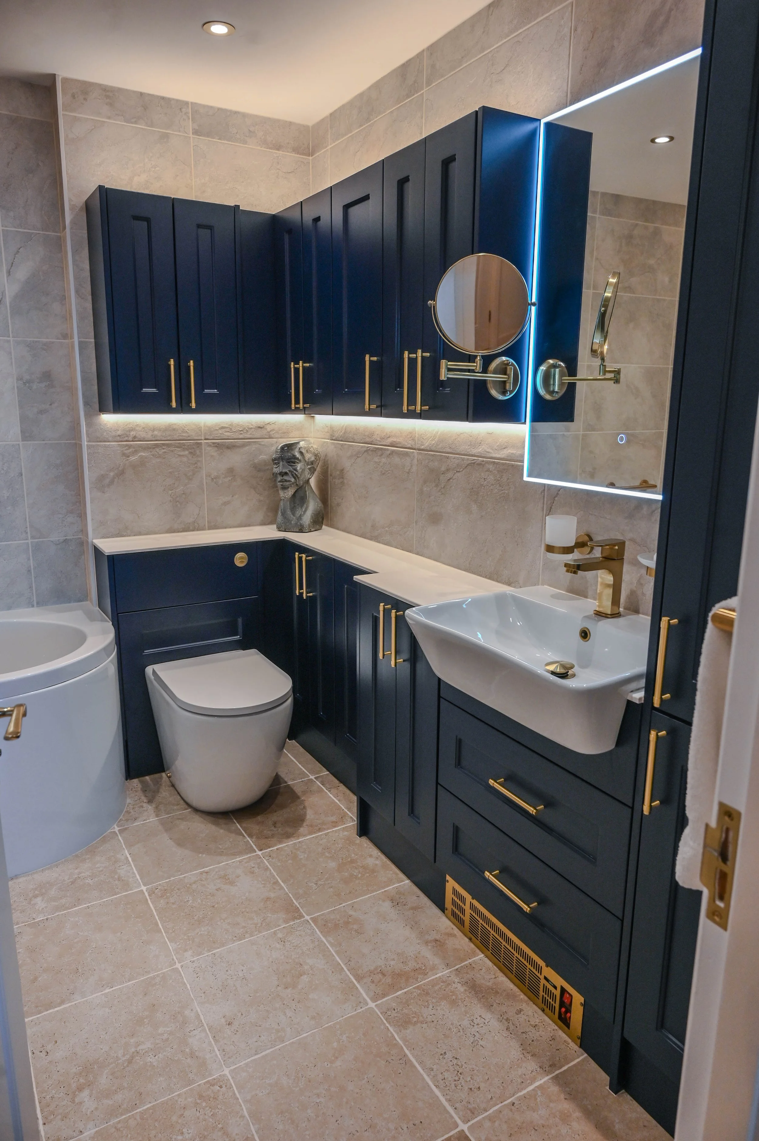 Modern bathroom with blue cabinetry, gold hardware, and beige tiled walls and floor. Includes a toilet, small sink with a gold faucet, a large mirror with LED lighting, a magnifying mirror, and decorative bust sculpture on the countertop.