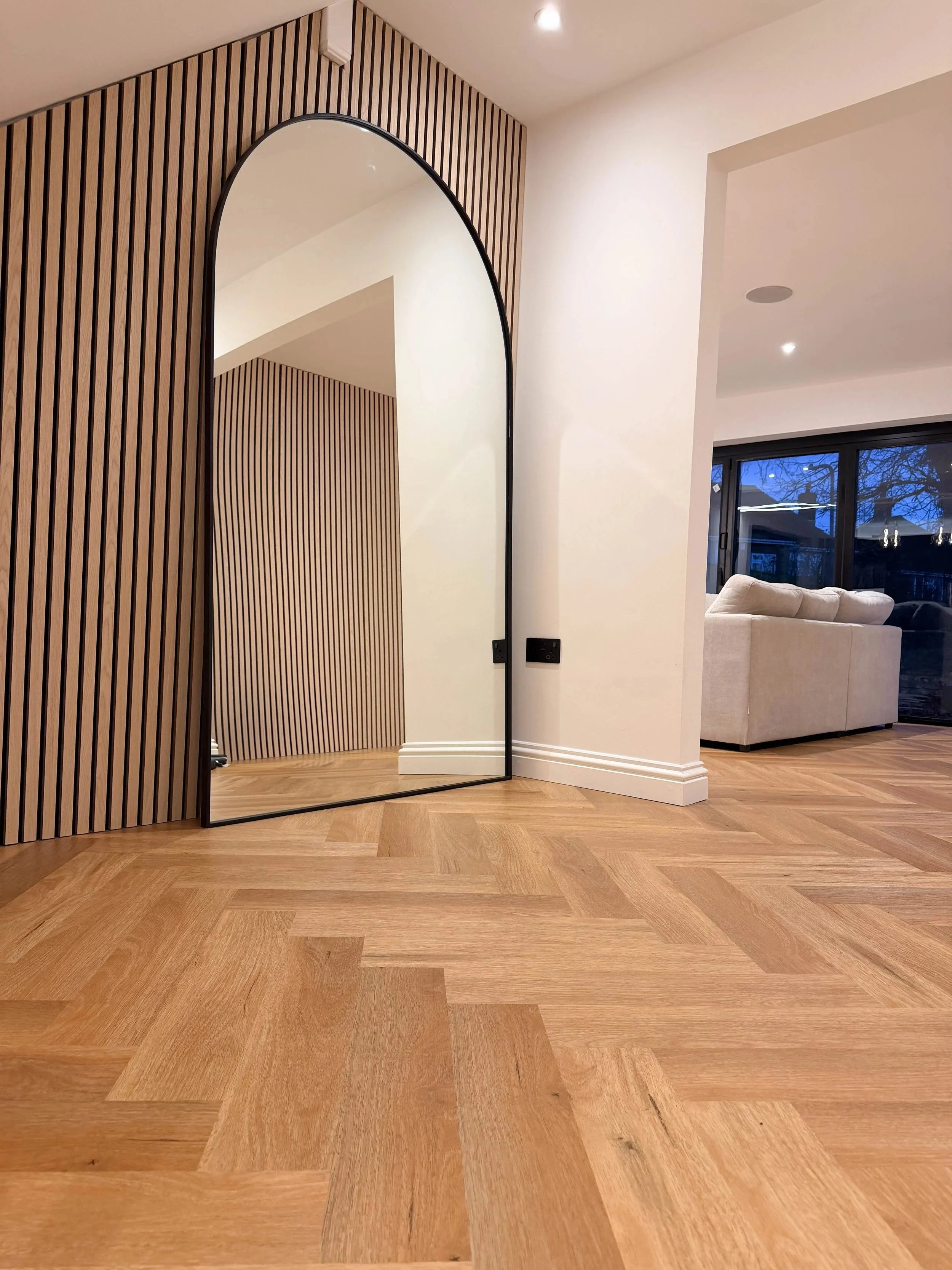 Modern interior with wooden flooring, a large arched mirror with a black frame, and a wall with vertical wooden slats. In the background, there is a living room with a white sofa and a large window showing trees outside at dusk.
