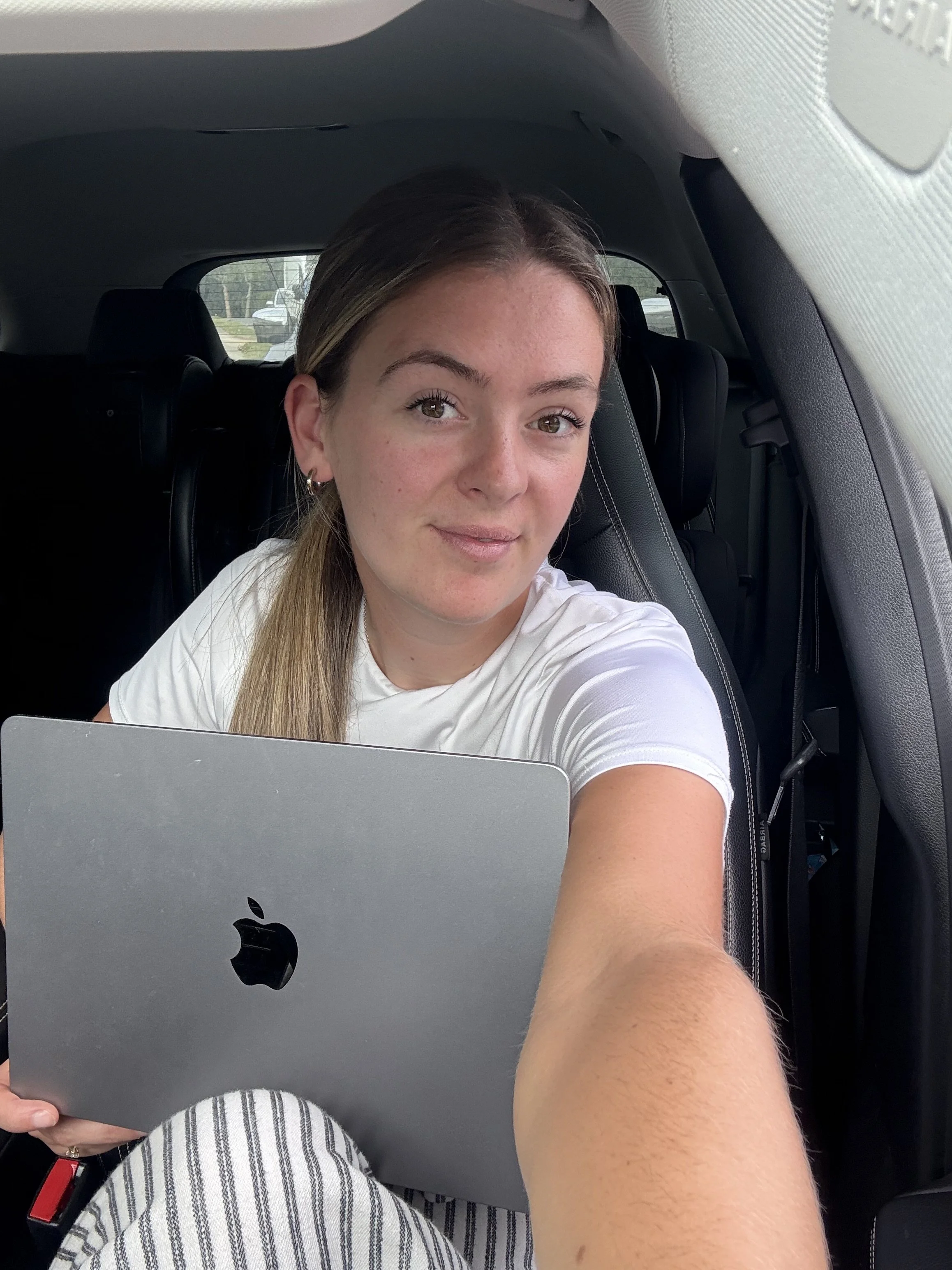 A young woman with long hair taking a selfie inside a car, holding a silver MacBook with the Apple logo visible, wearing a white t-shirt and striped pants.