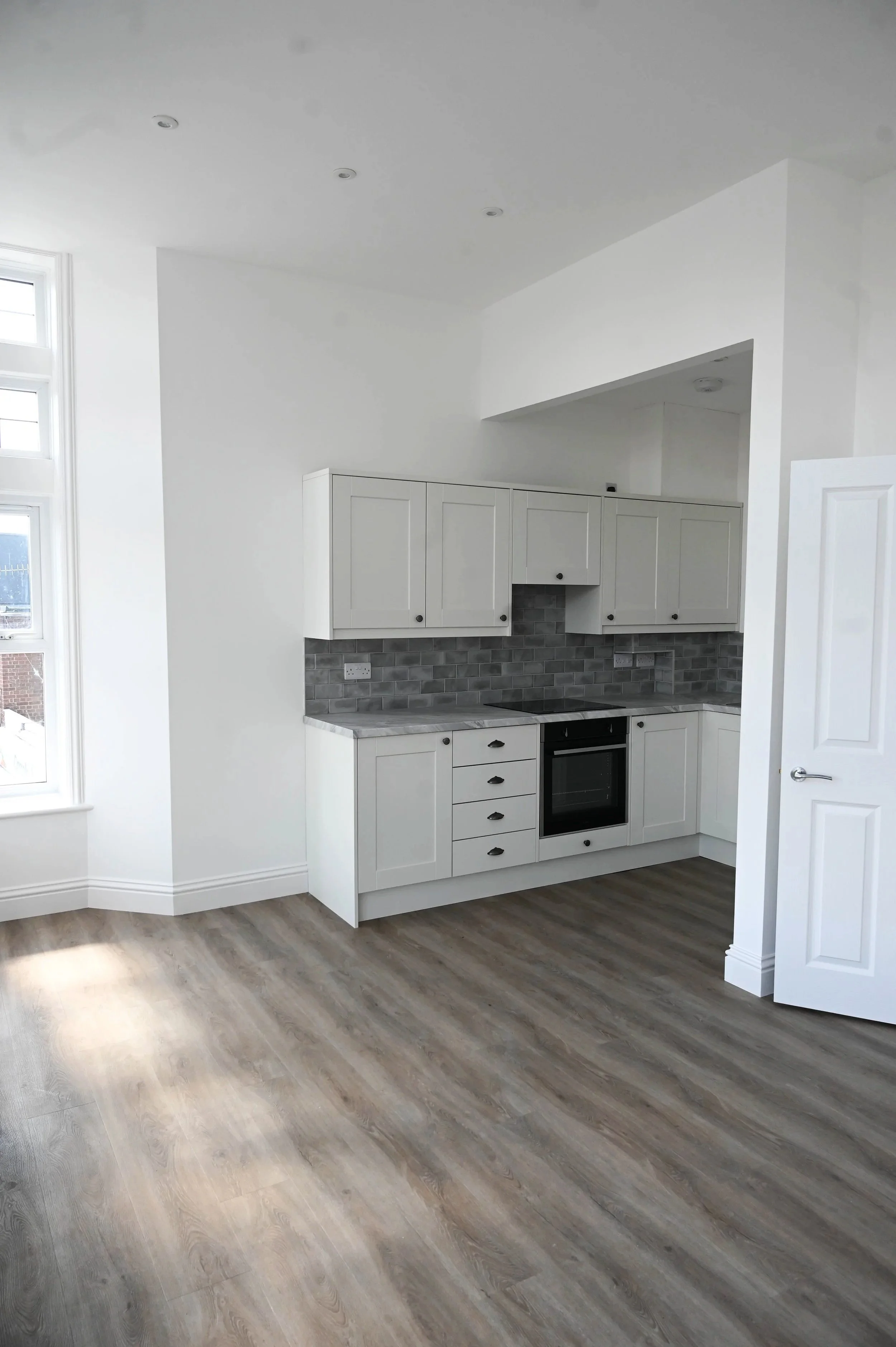 Empty kitchen with white cabinets, grey backsplash, black stove, and wood flooring.