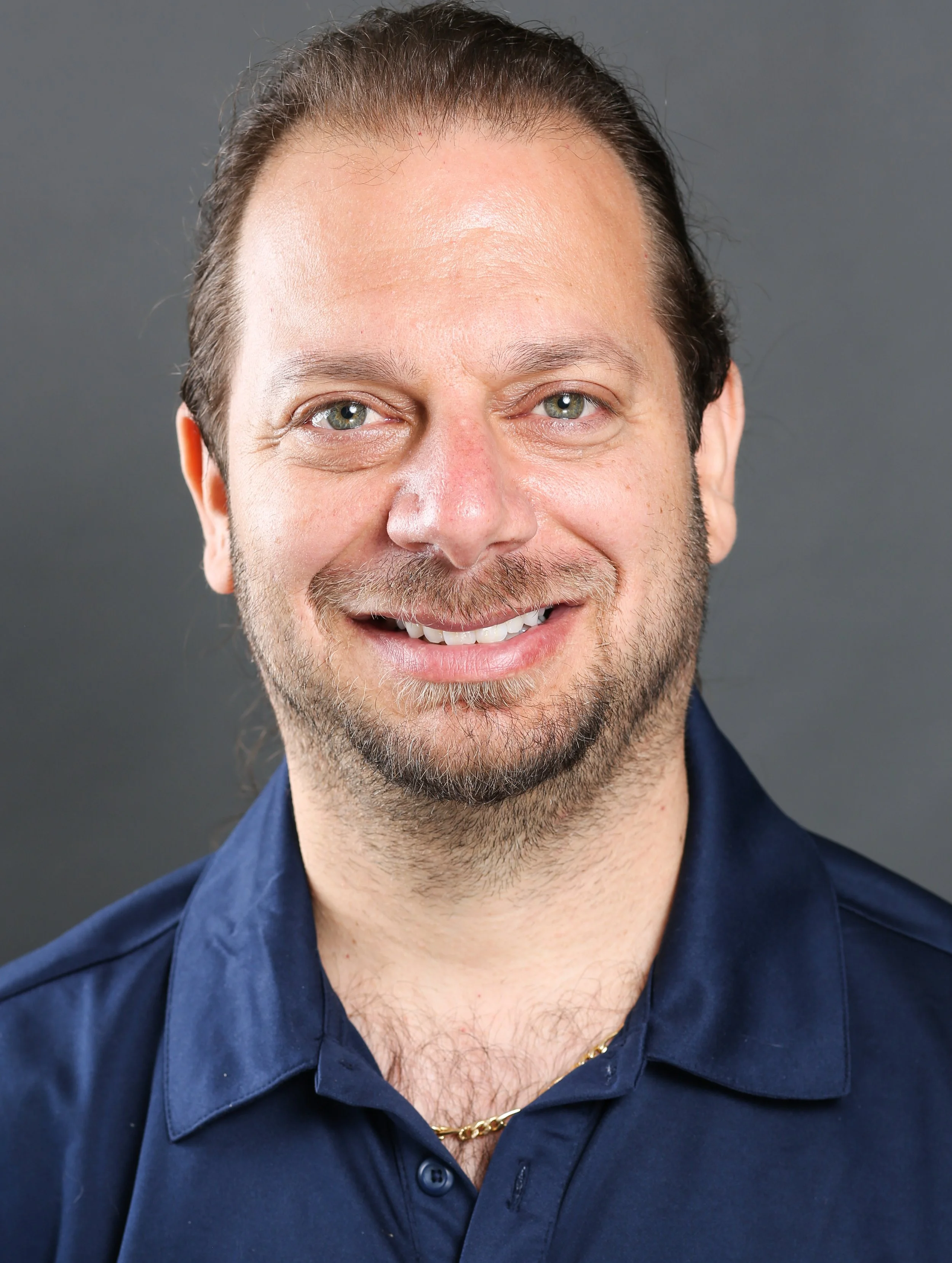 Close-up portrait of a smiling man with light skin, long brown hair, and a beard, wearing a dark blue collared shirt and a gold chain necklace, against a gray background.