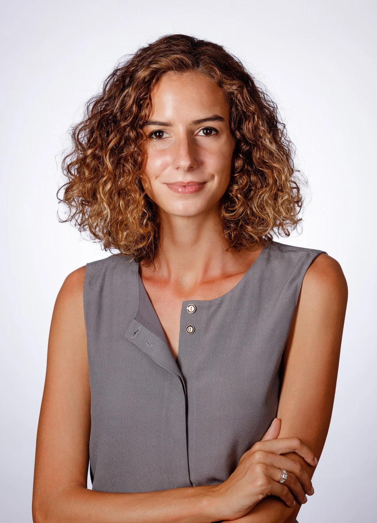 A woman with shoulder-length curly brown hair and light skin, wearing a sleeveless gray top, standing with her arms crossed against a plain white background.