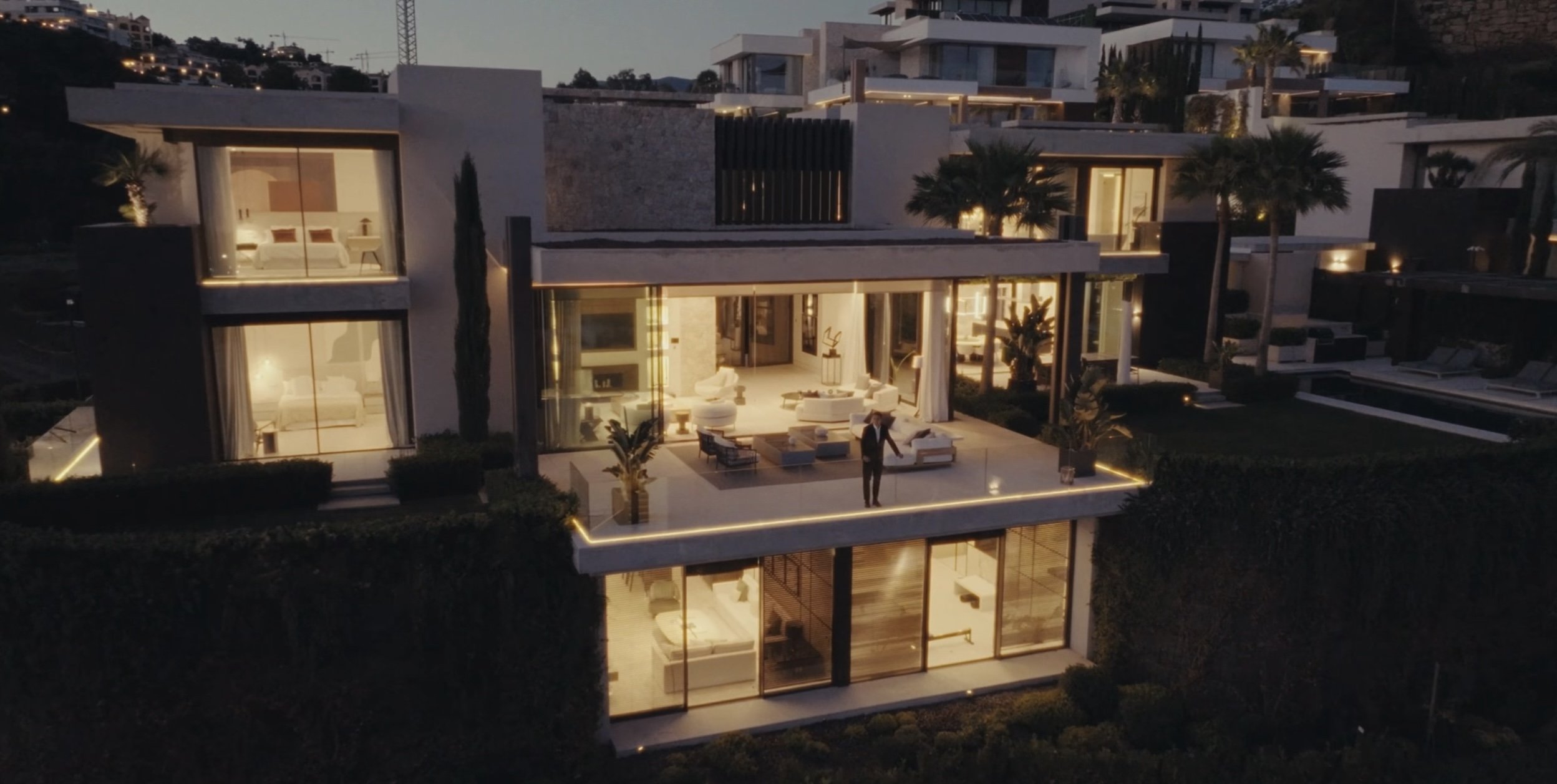 A modern multi-story house with large glass windows and balconies, illuminated at night. The house features outdoor seating on a spacious terrace with a person standing and talking outside. The interior visible through the windows shows stylish furniture and warm lighting, with a bedroom on the upper floors and a living room on the lower level.