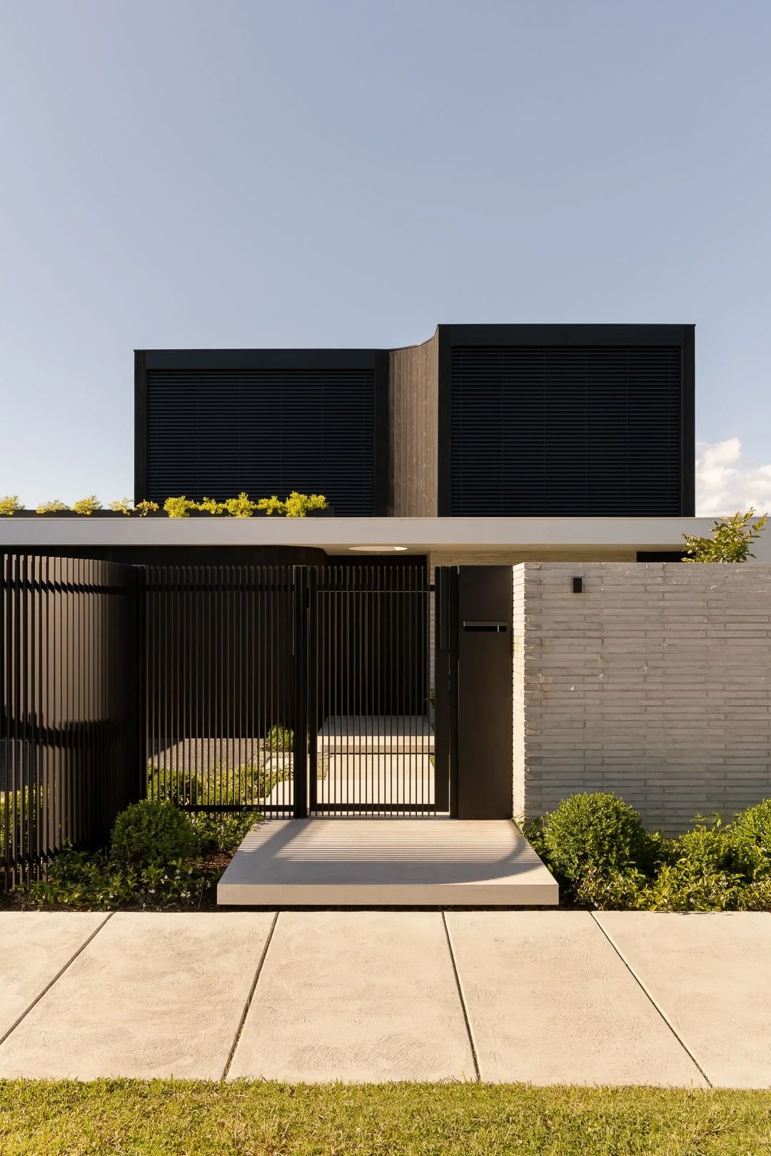 Modern house with black and gray exterior, black vertical slat fence, and small landscaped garden with bushes, under a clear blue sky.