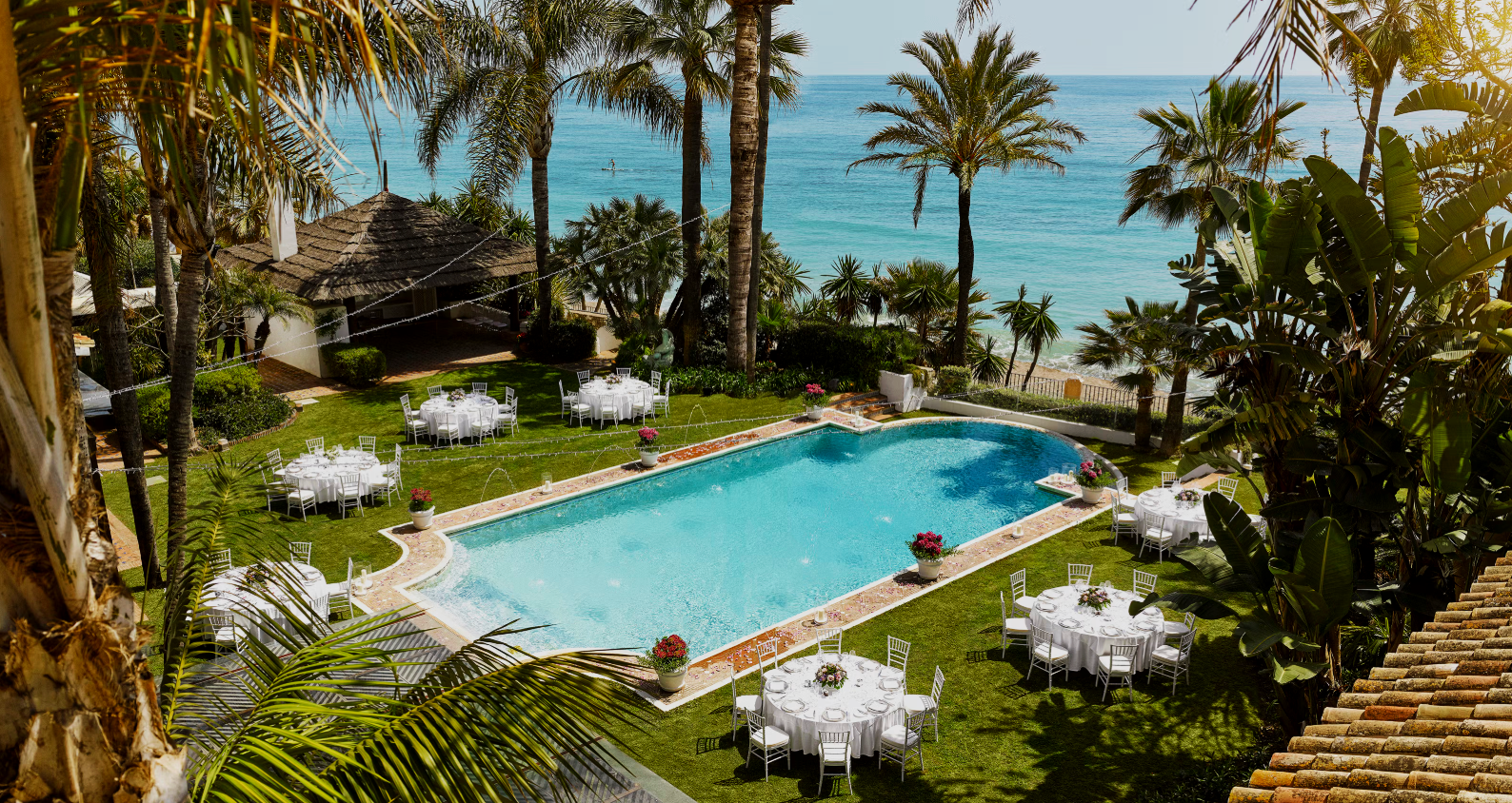 A scenic outdoor pool area with white-clothed round tables and chairs, surrounded by palm trees and overlooking the ocean. The setting appears to be prepared for a special event or gathering.