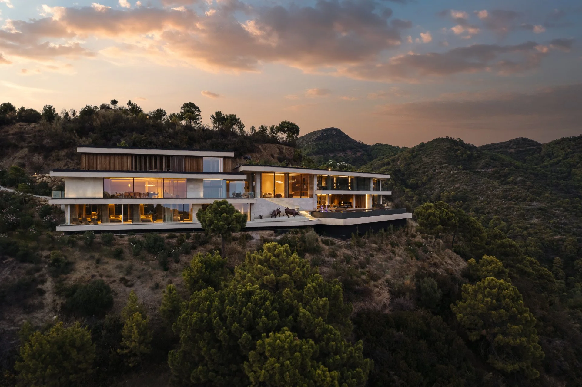 Modern multi-story house built on a hillside with large glass windows and spacious balconies, surrounded by trees and hills, during sunset.