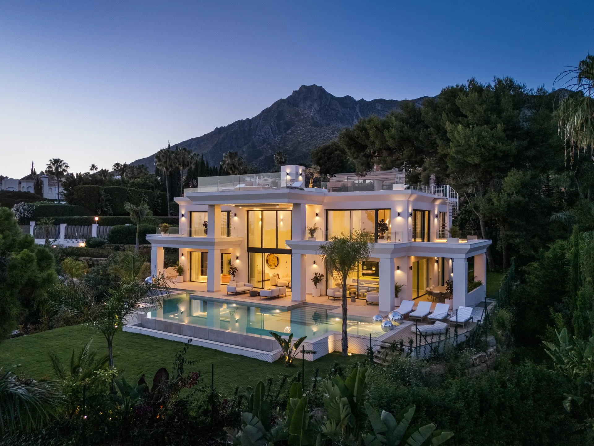 Luxury modern house with large glass windows, outdoor pool, and outdoor seating, surrounded by lush greenery and mountains at dusk.