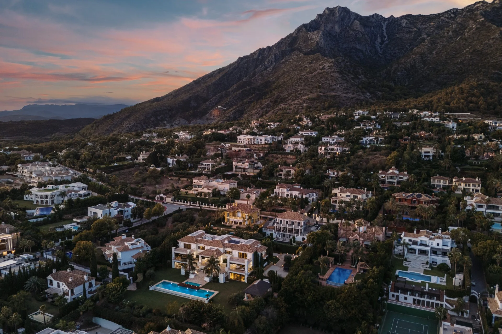 Aerial view of a hillside residential area with luxurious white and yellow villas, swimming pools, tennis courts, lush greenery, and mountain backdrop during dusk.