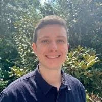 Smiling person outdoors with trees and greenery in the background.