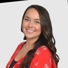 Woman smiling, wearing a red floral top with long brown hair against a light gray background.