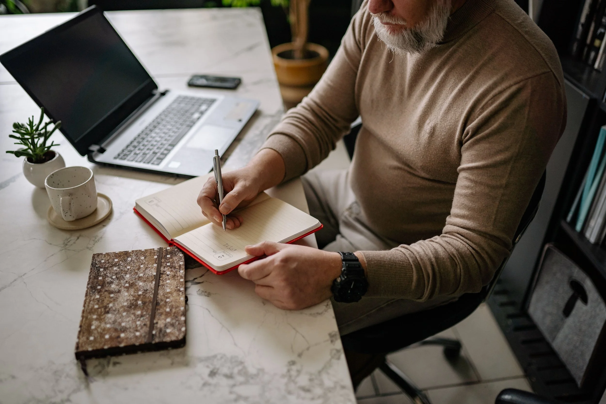 older man at a desk with a laptop holding a pencile over a notebook