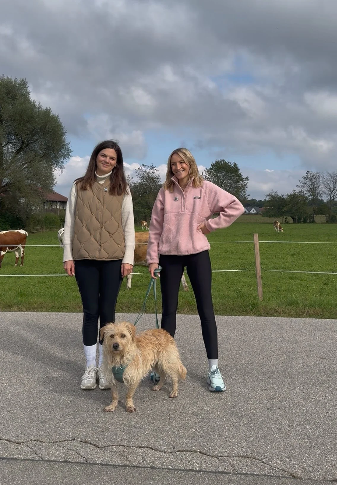 Zwei junge Frauen mit einem Hund auf einer Straße, im Hintergrund eine grüne Wiesenlandschaft mit einigen Kühen und Bäumen, bewölkter Himmel.