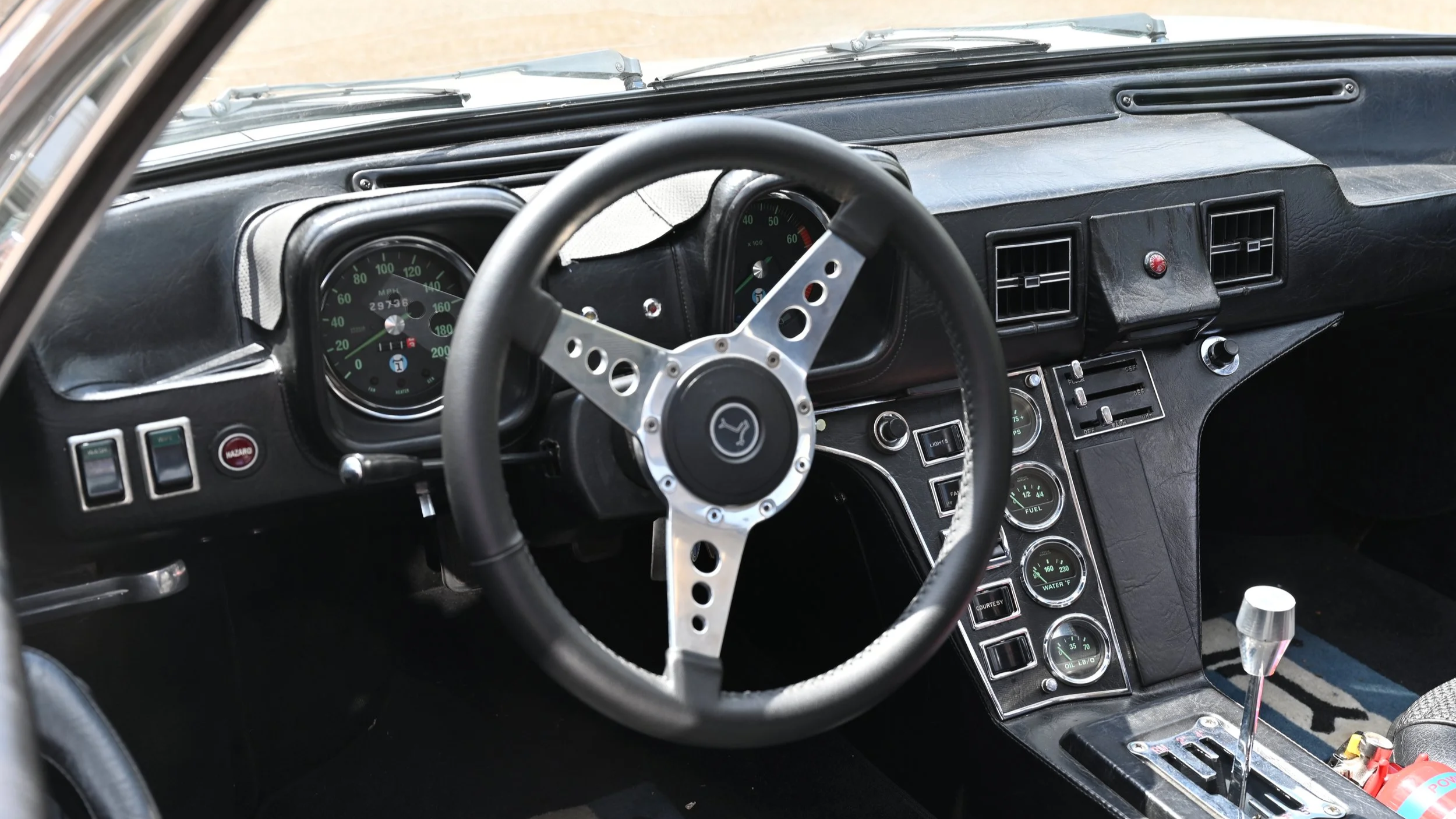 Interior of a vintage car showing a black dashboard with analog gauges, a steering wheel with a racing style design, and a gear shift lever.