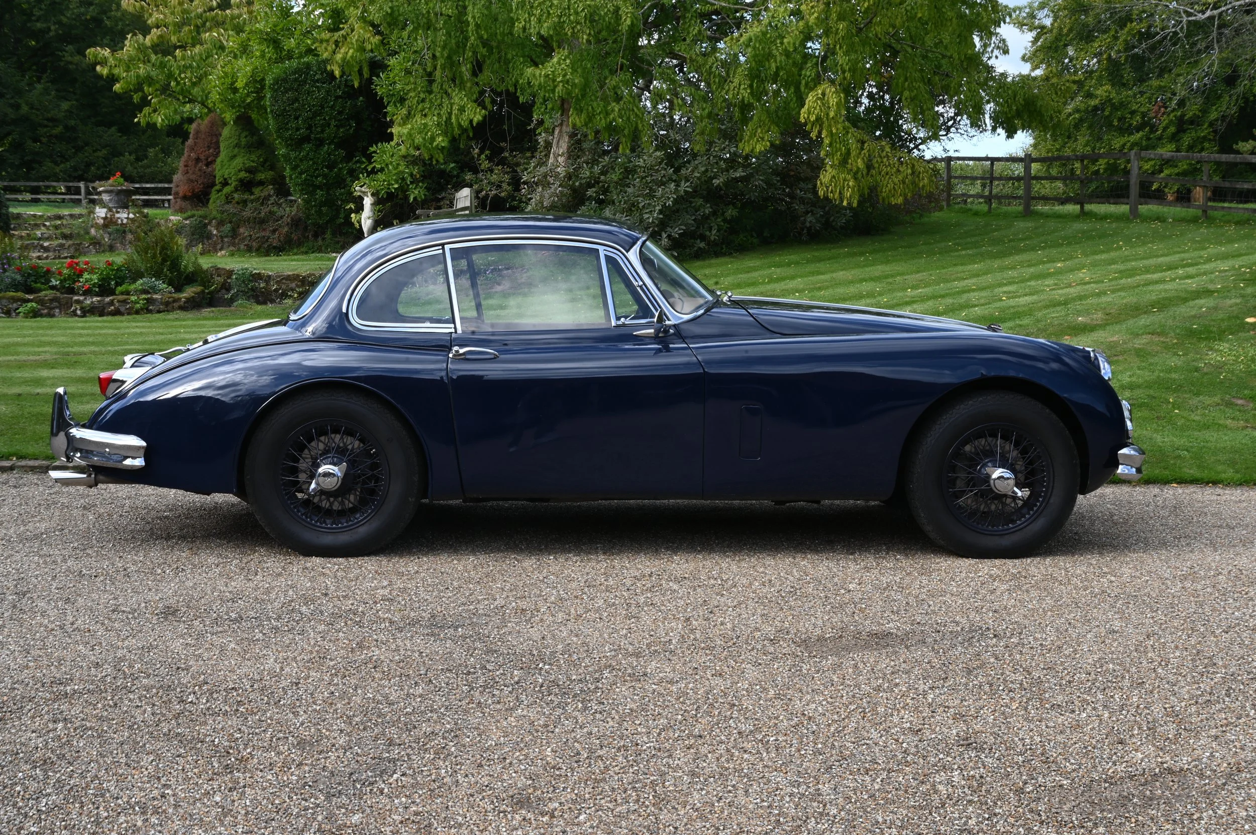 A vintage navy blue sports car with wire-spoked wheels parked on a gravel driveway in a lush green garden setting.