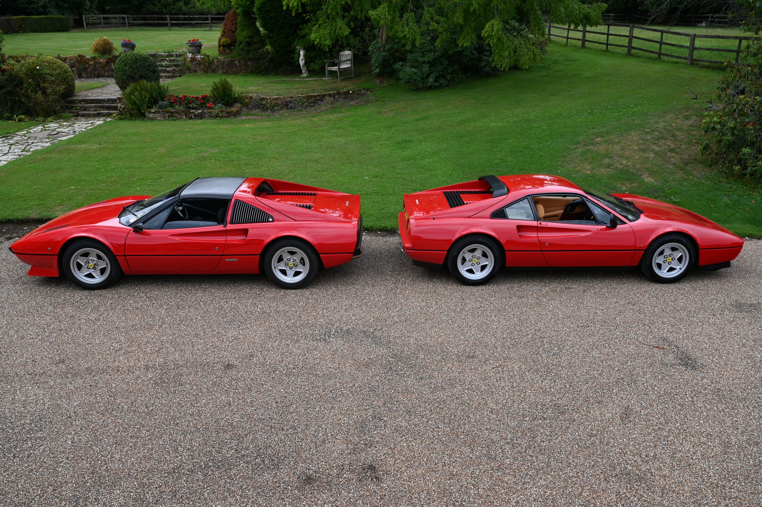 Two red Ferrari sports cars parked side by side on a driveway in front of a well-kept garden with green lawns, bushes, and trees.