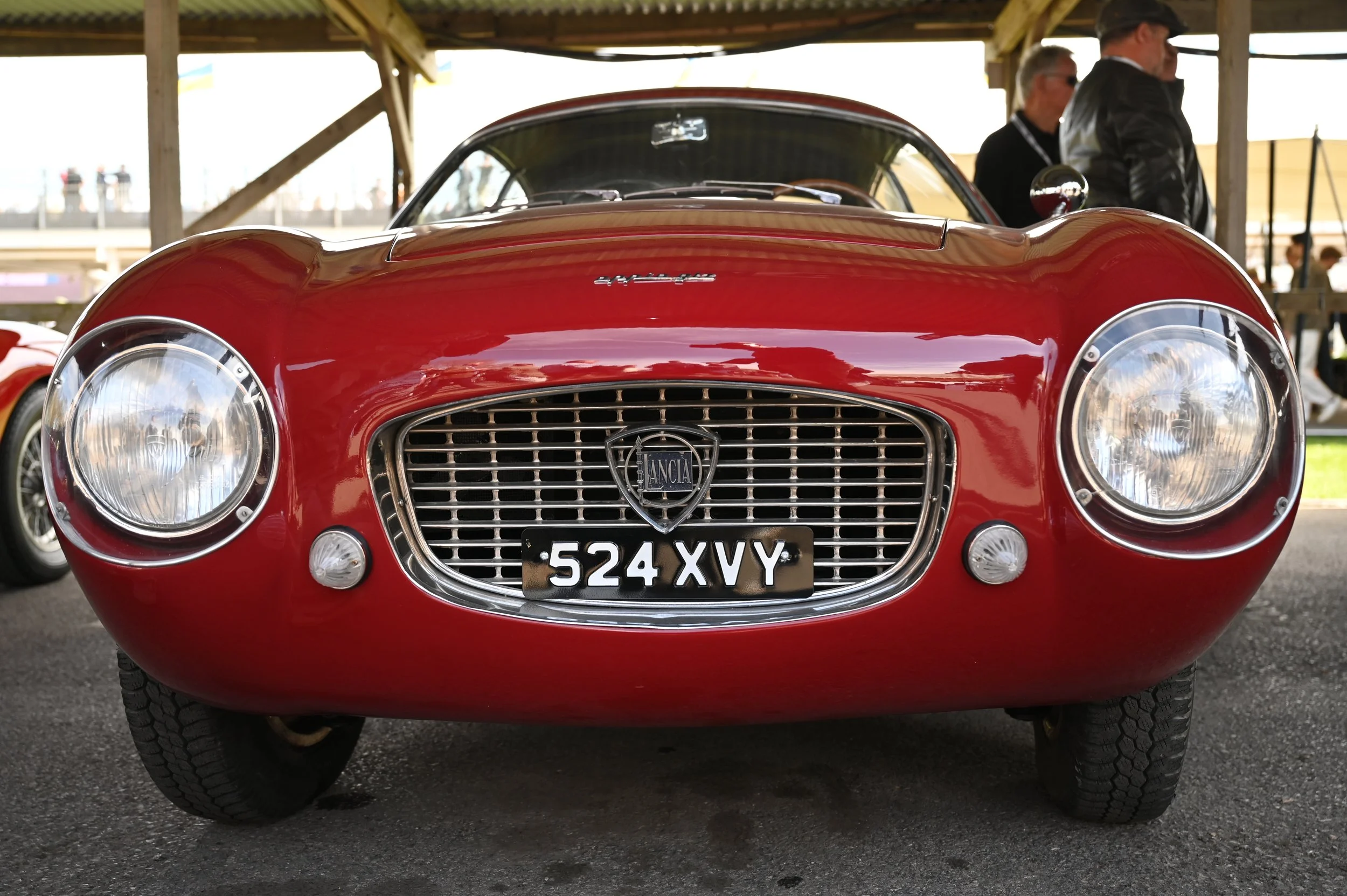 Front view of a red vintage Lancia sports car with a front grille and round headlights at an outdoor car show, with people in the background.