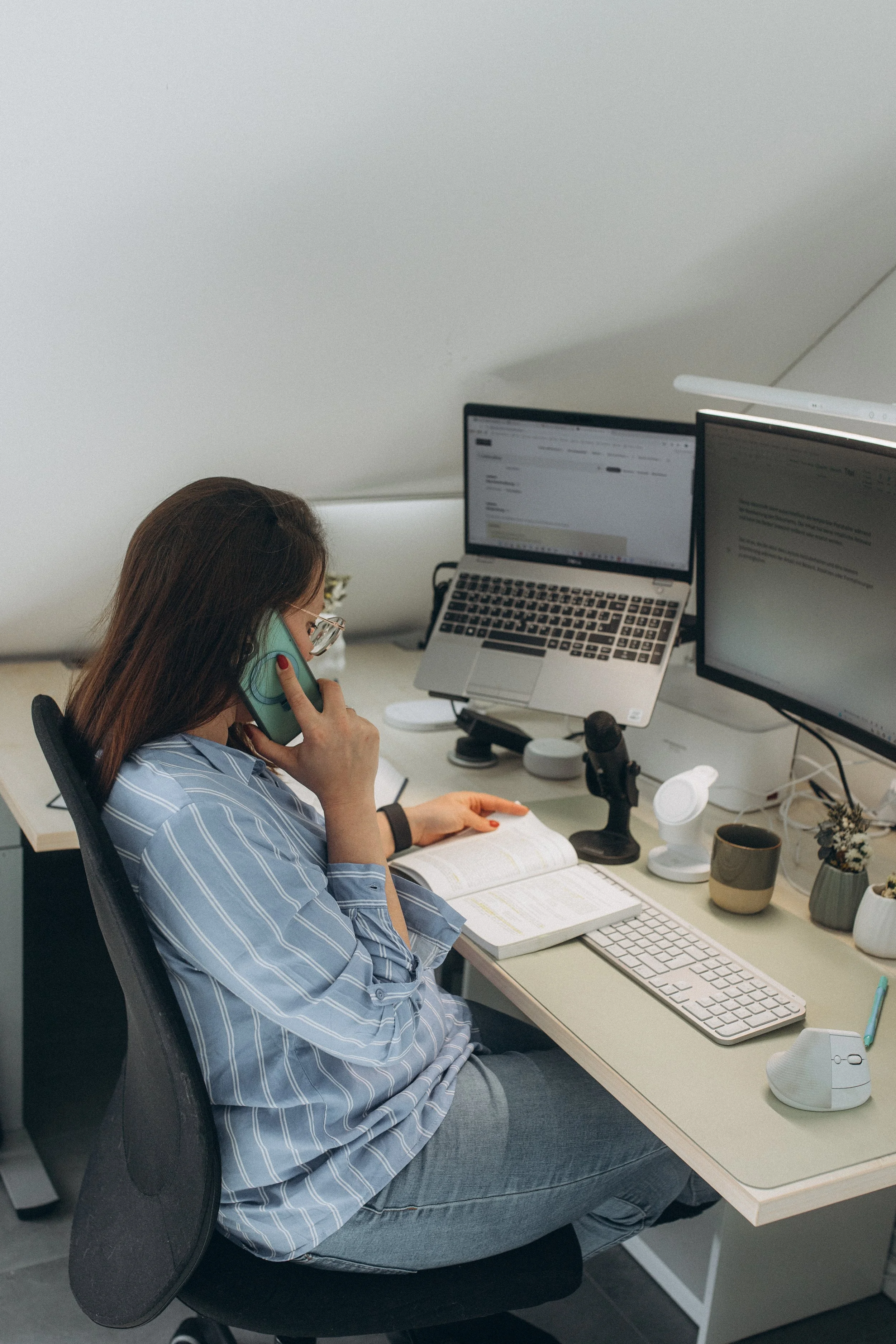 Eine Frau sitzt an einem Schreibtisch, spricht am Telefon und arbeitet an zwei Computerbildschirmen, mit Büchern und Schreibutensilien auf dem Tisch.