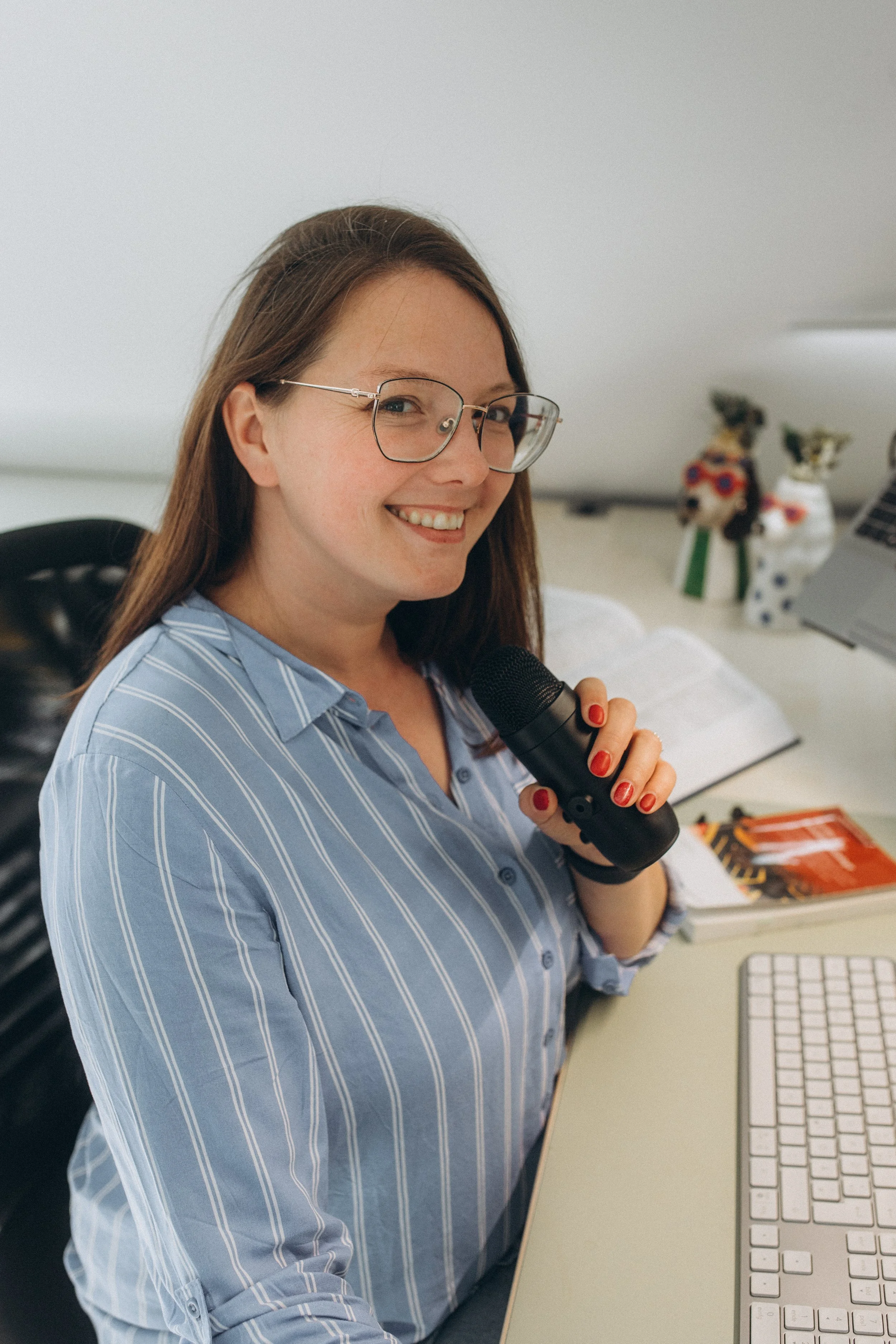 Eine lachende Frau mit Brille hält ein Mikrofon in der Hand, vor einem Laptop auf einem Schreibtisch, im Hintergrund sind dekorative Katzenfiguren.