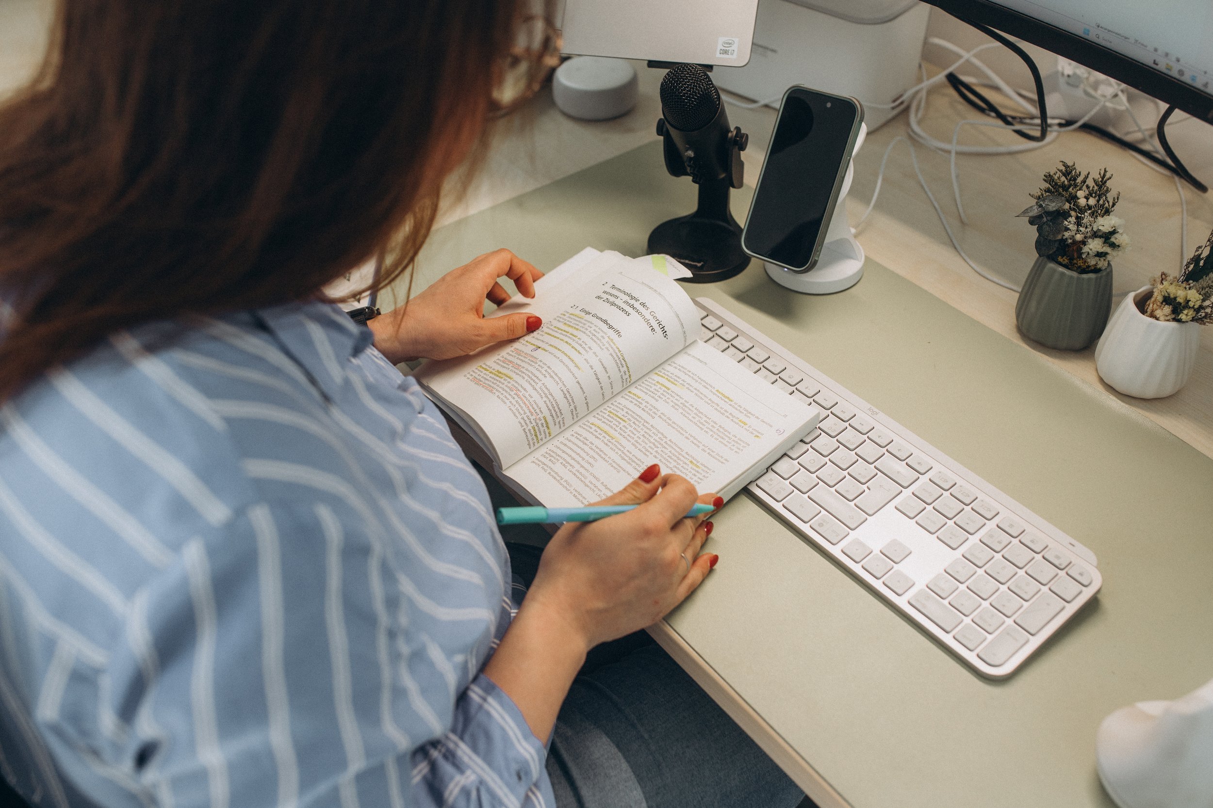 Eine Person liest in einem Buch, während sie an einem Schreibtisch sitzt. Auf dem Schreibtisch befinden sich eine Tastatur, ein Monitor, ein Smartphone, ein Mikrofon, ein Notizblock und ein Kugelschreiber. Im Hintergrund sind kleine Pflanzen auf dem Tisch.