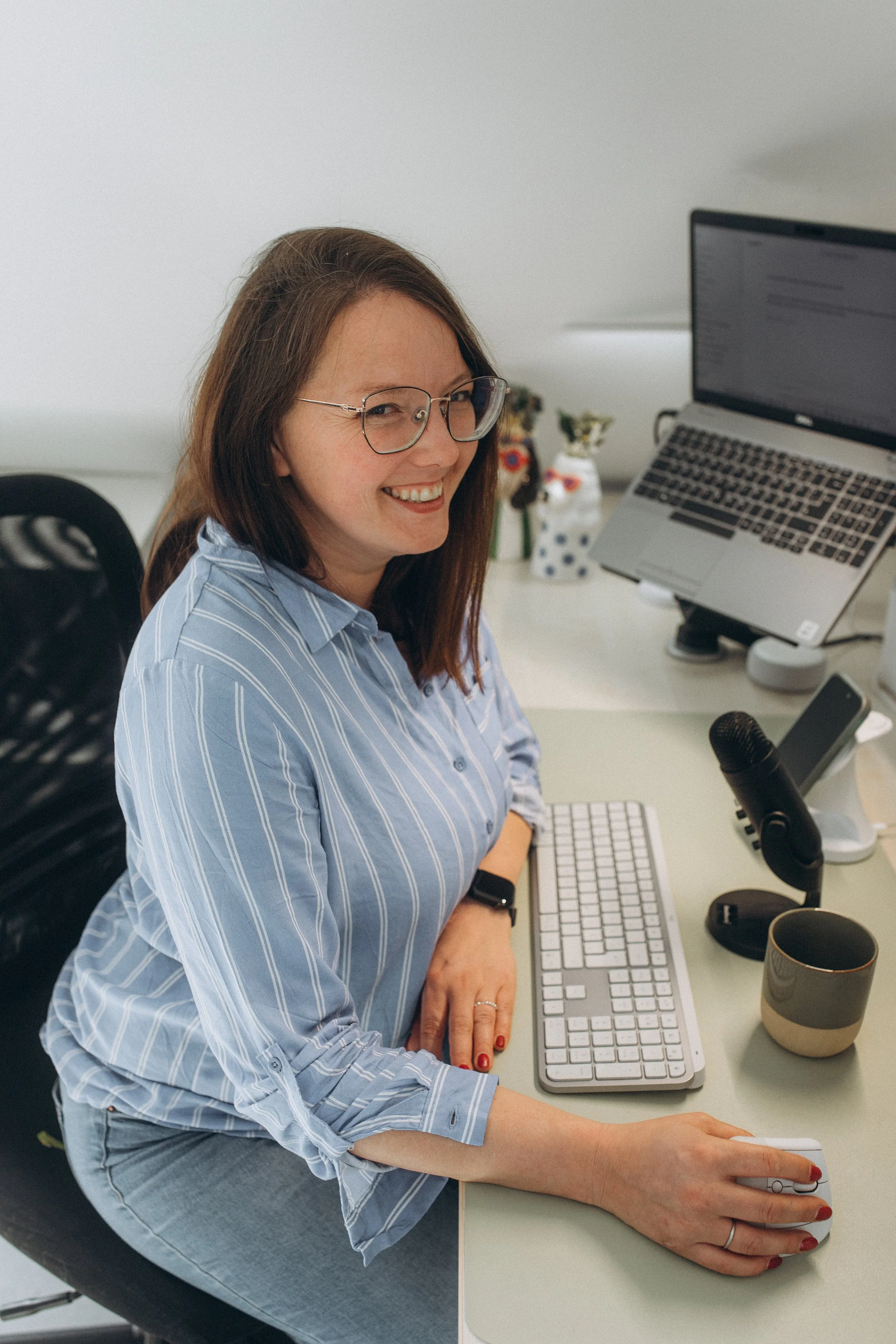 Eine lachende Frau mit Brille sitzt an einem Schreibtisch mit Computer, Tastatur, Mikrofon und Getränk, im Hintergrund befinden sich Dekorationsgegenstände.