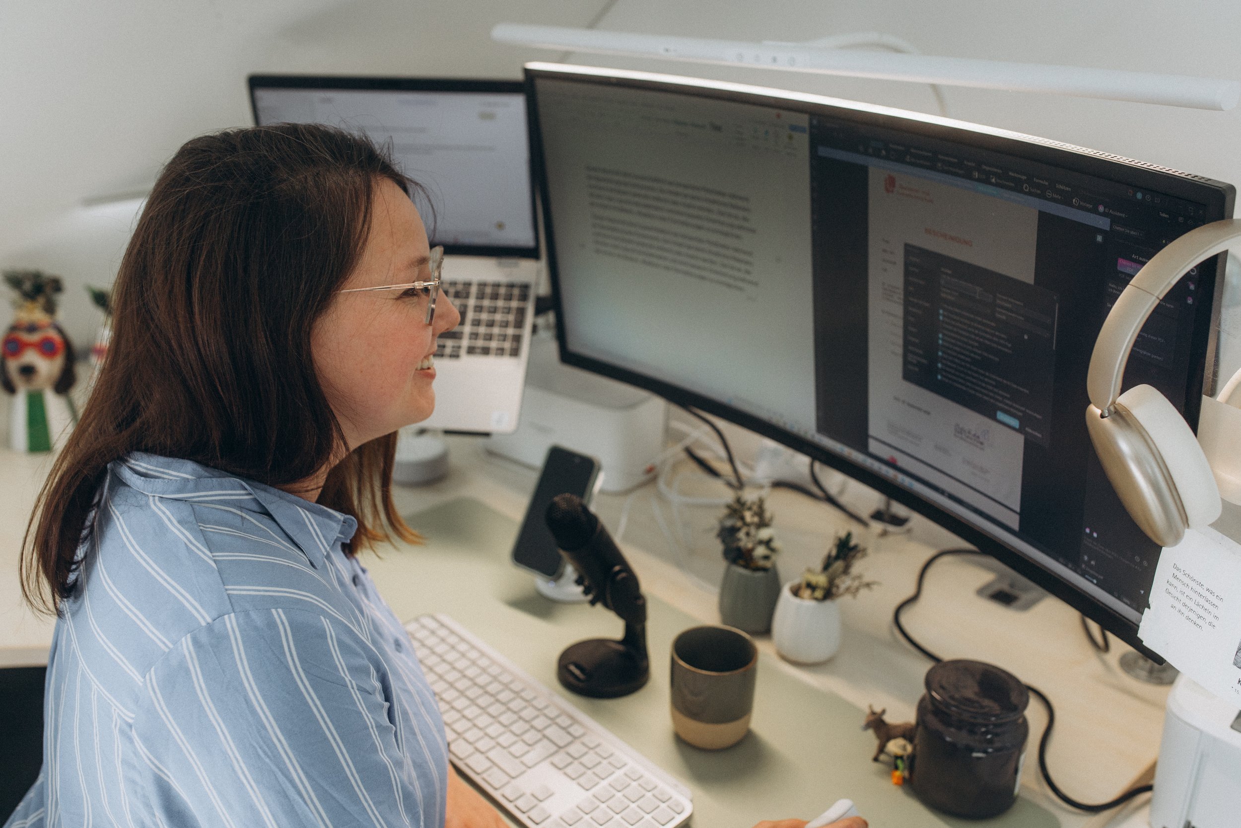 Frau arbeitet an einem Schreibtisch mit zwei Computermonitoren, Tastatur, Maus und weiteren Büromaterialien, im Hintergrund Dekorationen und Pflanzen.