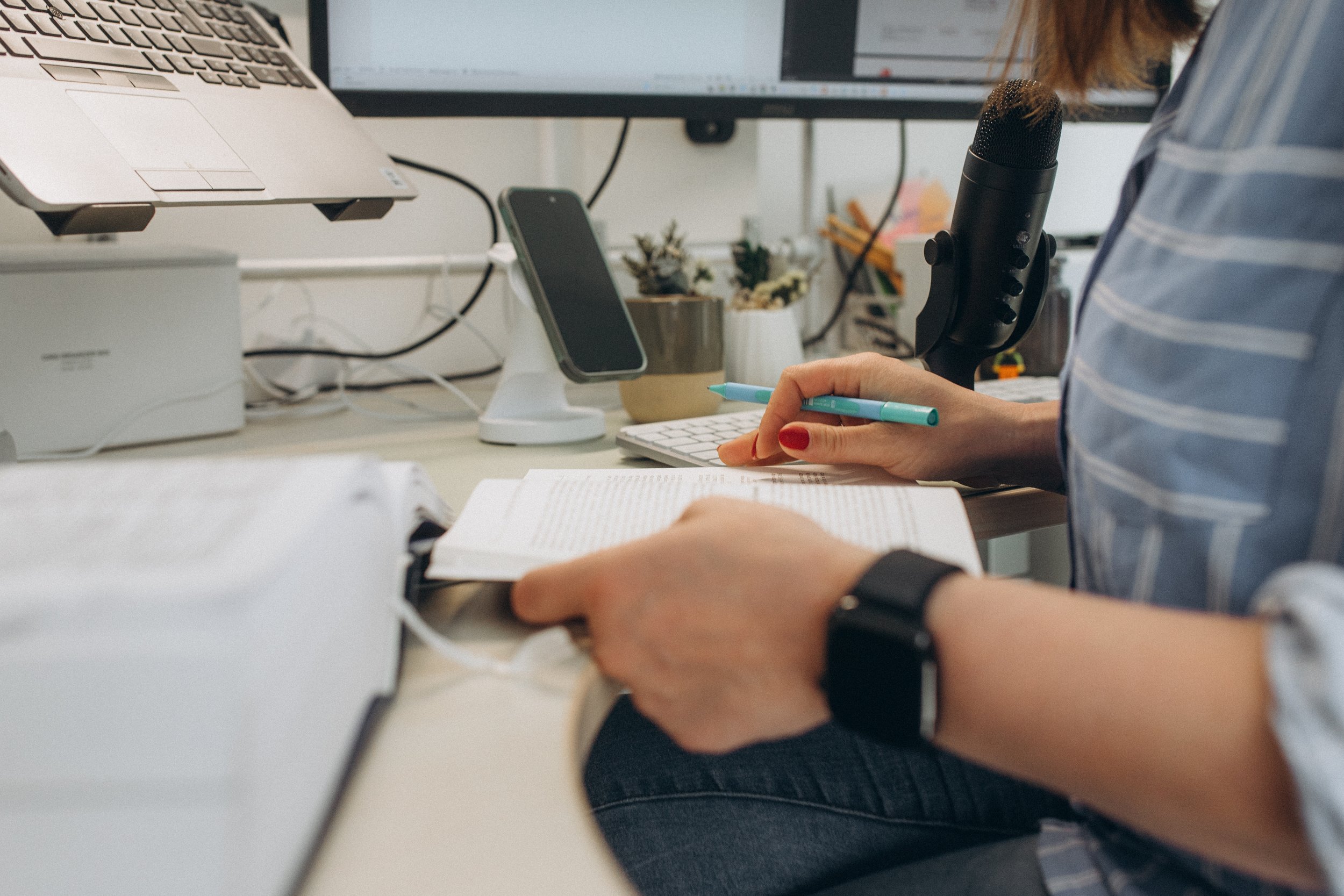 Eine Person arbeitet an einem Schreibtisch mit einem Laptop, einem Smartphone auf einem Ständer, einem Mikrofon, einem Keyboard und verschiedenen Pflanzen im Hintergrund.