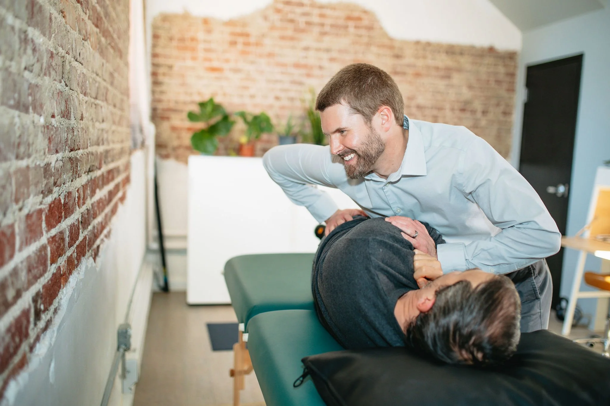 A chiropractor adjusting a male patient lying on his side on an examination table, smiling in a well-lit room with brick walls and houseplants.
