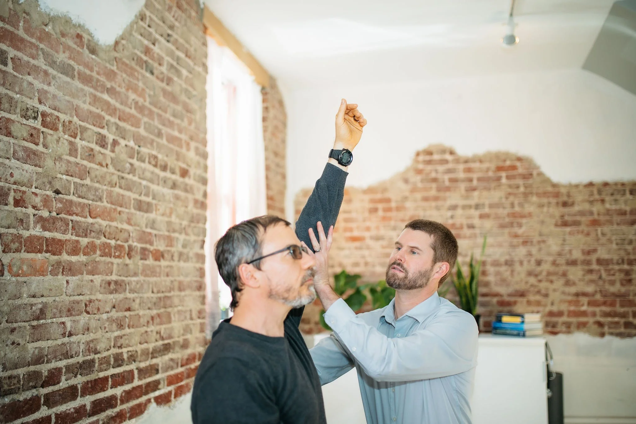Man in a gray shirt having his arm measured or examined by another man in a black shirt in a room with exposed brick walls.