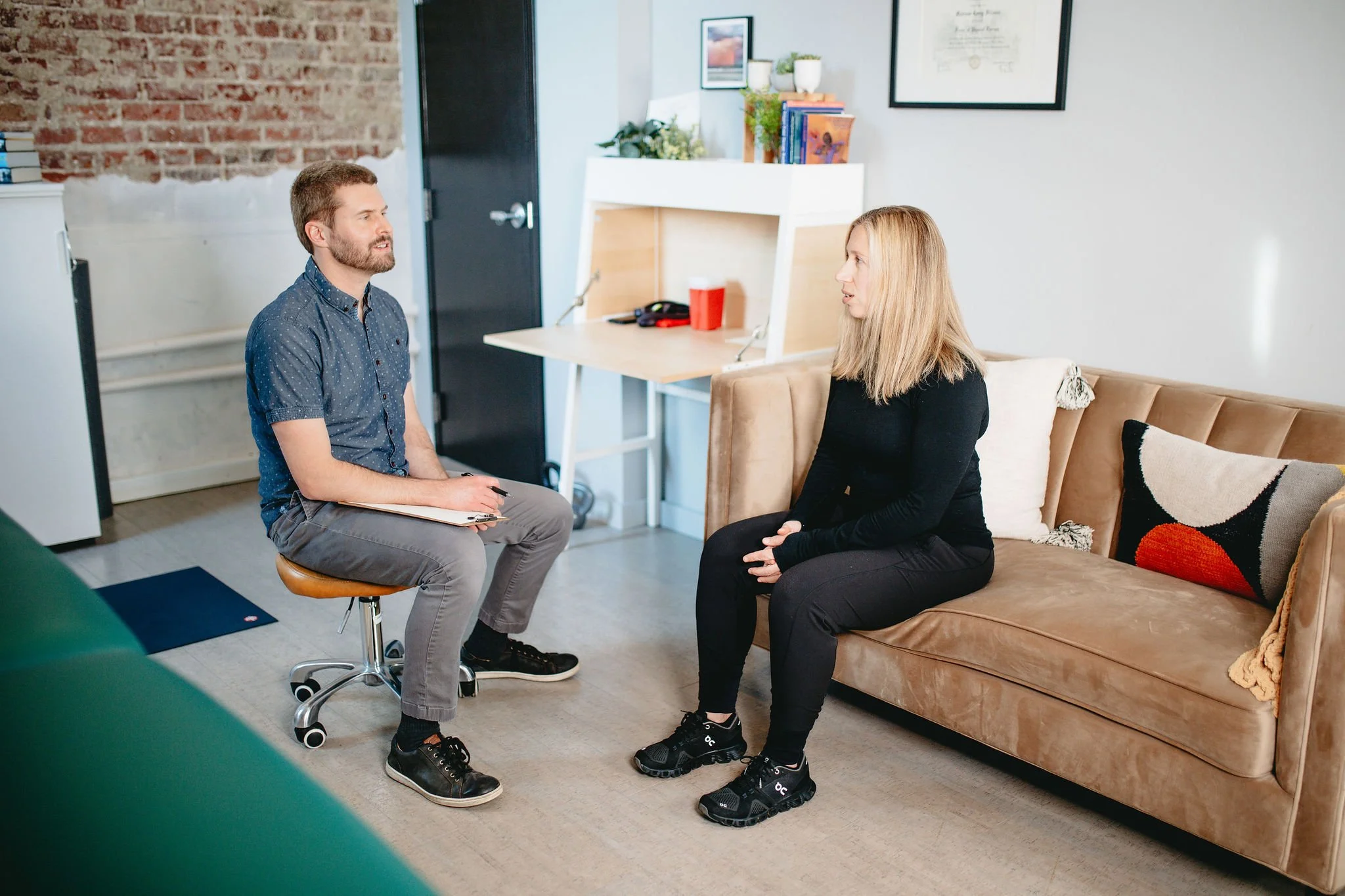 A therapist sitting on a stool and talking to a client who is sitting on a beige sofa in a therapy office. The room has a brick wall, a small desk, a bookshelf, and framed artwork on the wall.