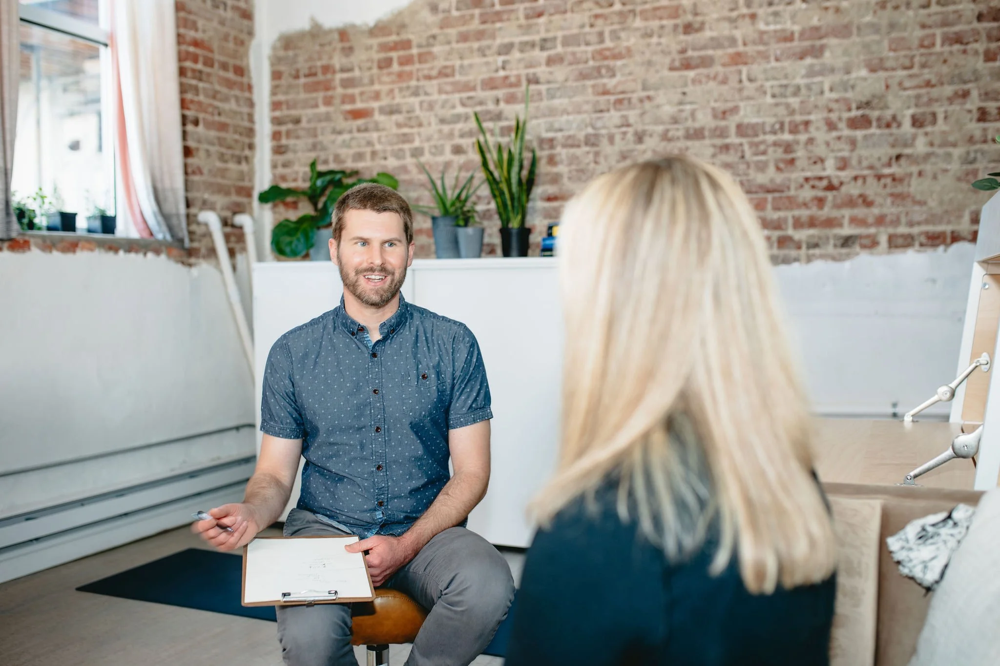 A man with a clipboard talking to a woman during a counseling or interview session in a bright room with a brick wall and potted plants.