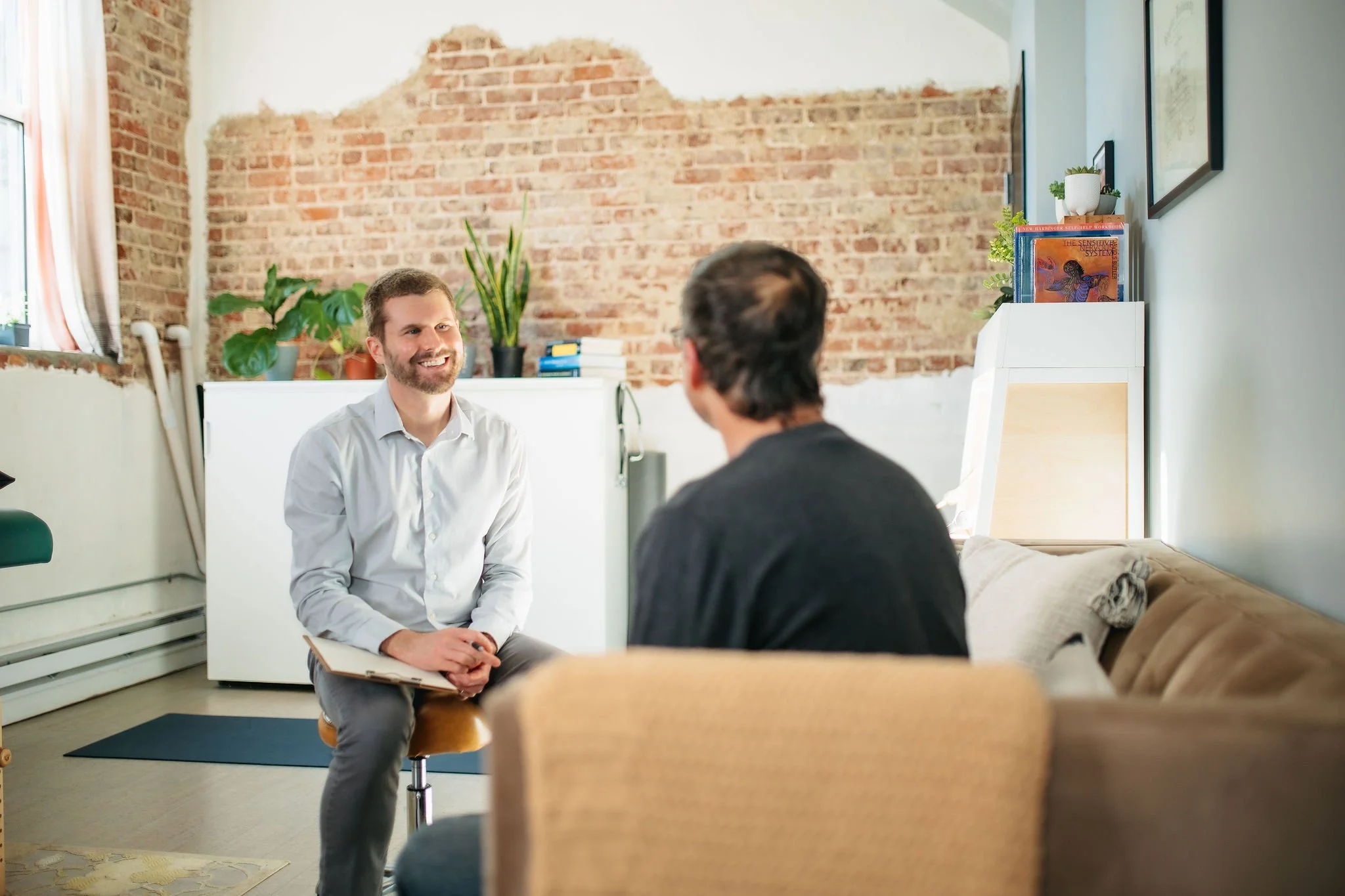 Two men sitting in an office, engaged in conversation. One man, facing the camera, is smiling and holding a notebook. The other man has his back to the camera. The room has a brick wall, a window with curtains, and potted plants.