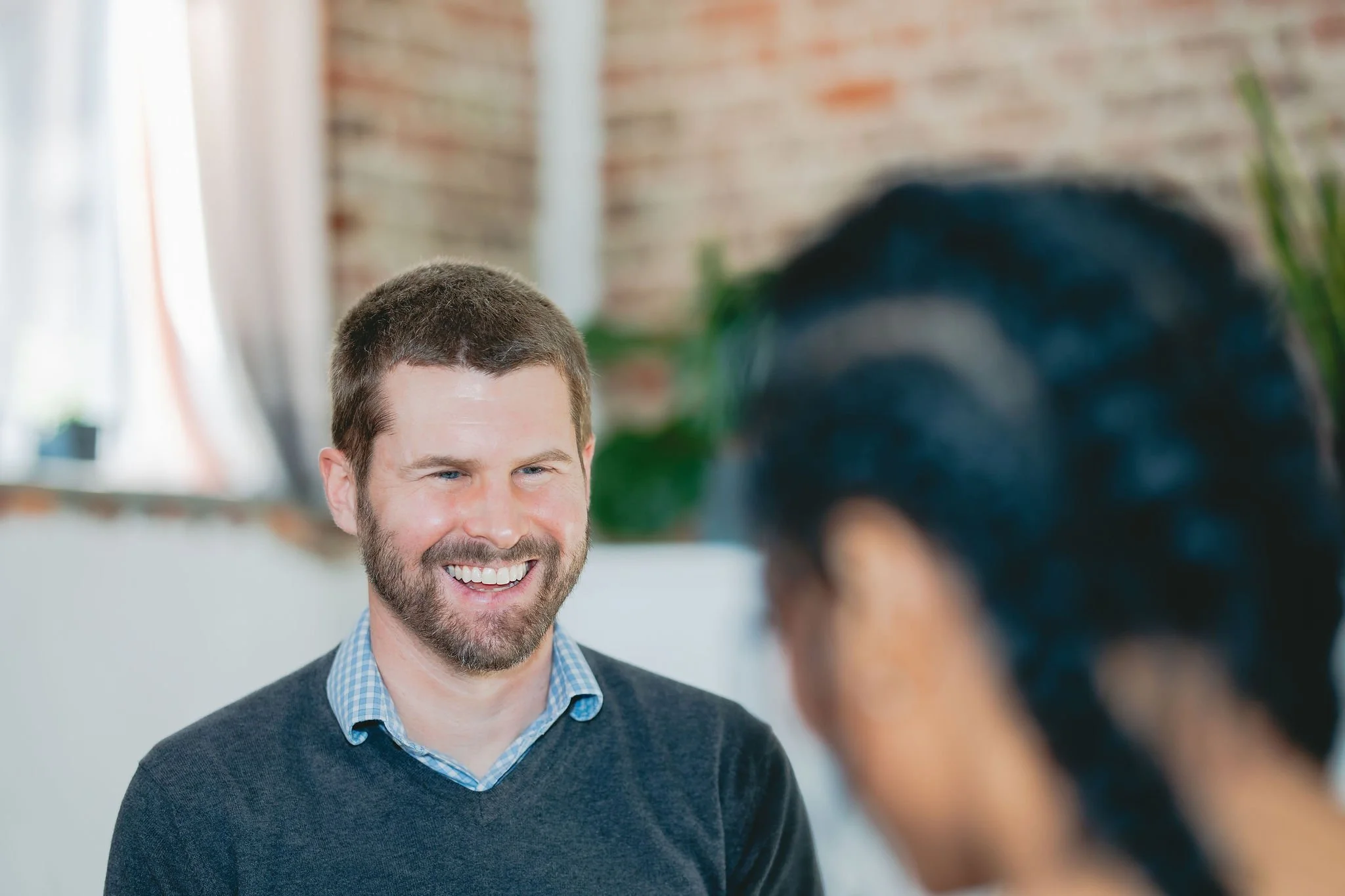 A man with short brown hair, beard, and blue eyes smiling while talking to a woman with black hair and glasses in an indoor setting with brick wall and curtains in the background.