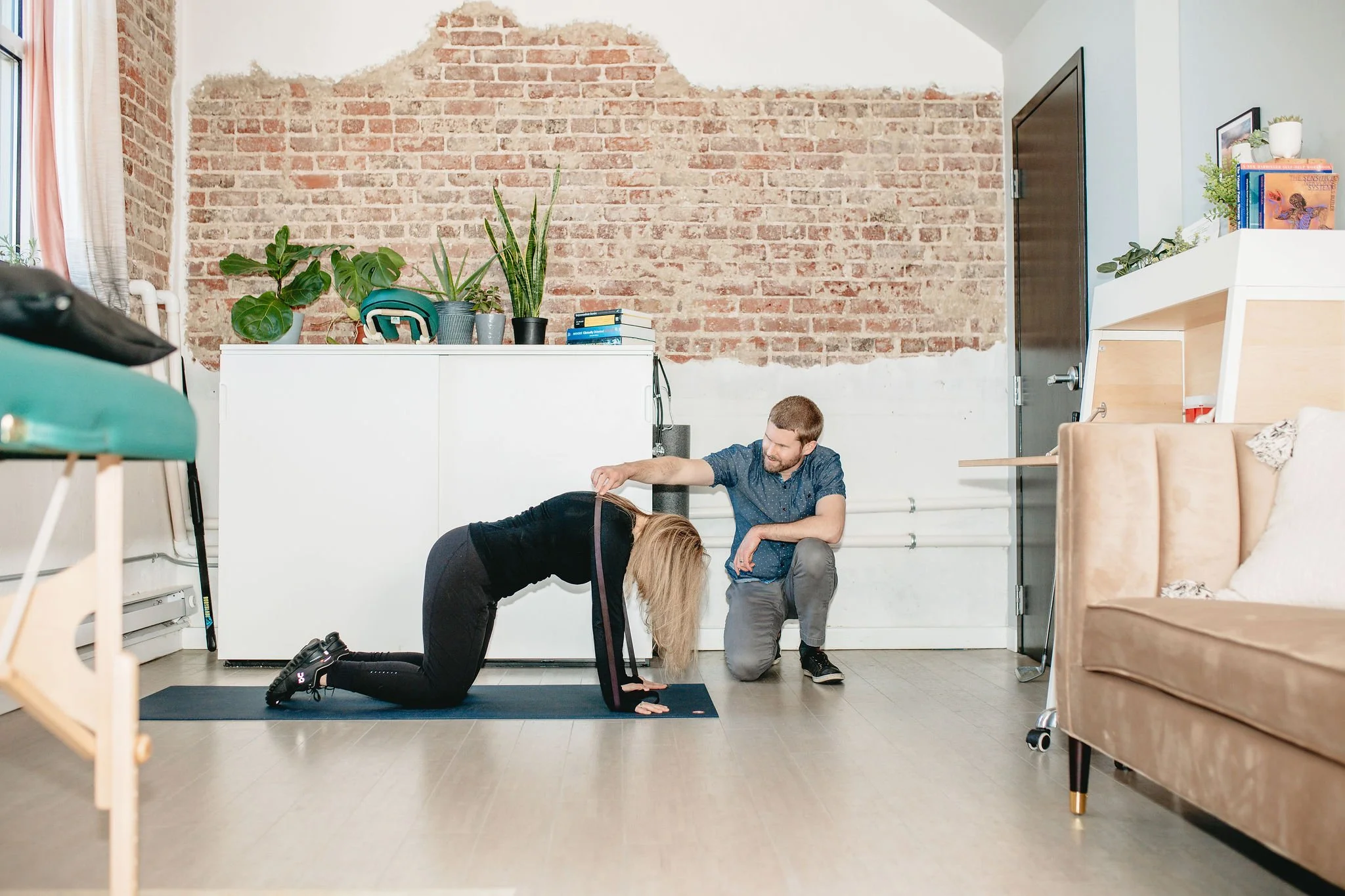 A woman performing a plank exercise on a yoga mat while a man offers encouragement with a measuring tape around her waist in a cozy, well-lit living room with exposed brick wall and houseplants.
