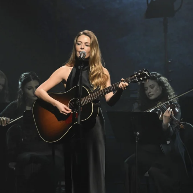 Maggie Rogers sings while strumming a guitar, looking stunning in a satiny black dress. Two women play violins in the background.