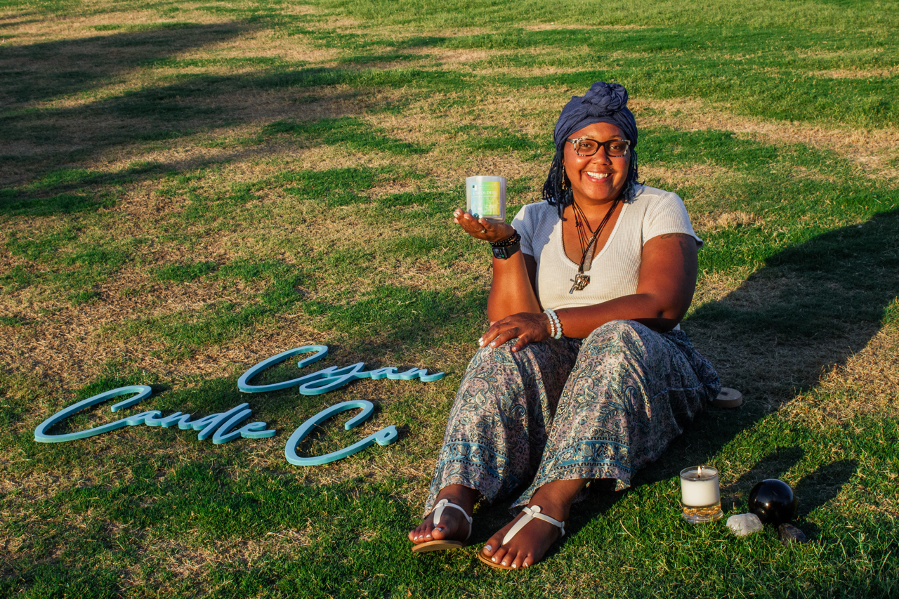 Mya from Cyan Candle Co. sitting on grass holding a candle with lit and unlit candles and decorative sign that says 'Can You C?