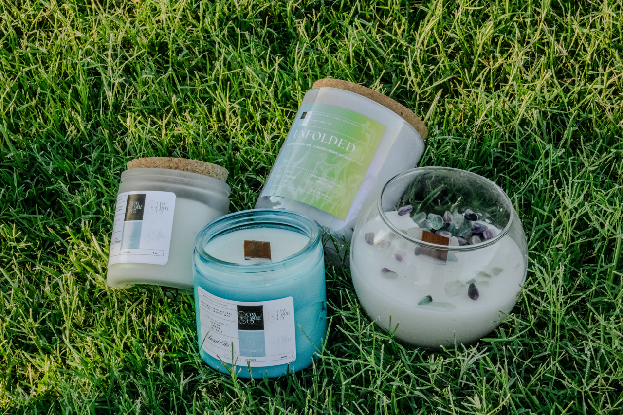 Mya from Cyan Candle Co. Four candles in glass and metal containers, one with purple stones and white wax, on green grass.