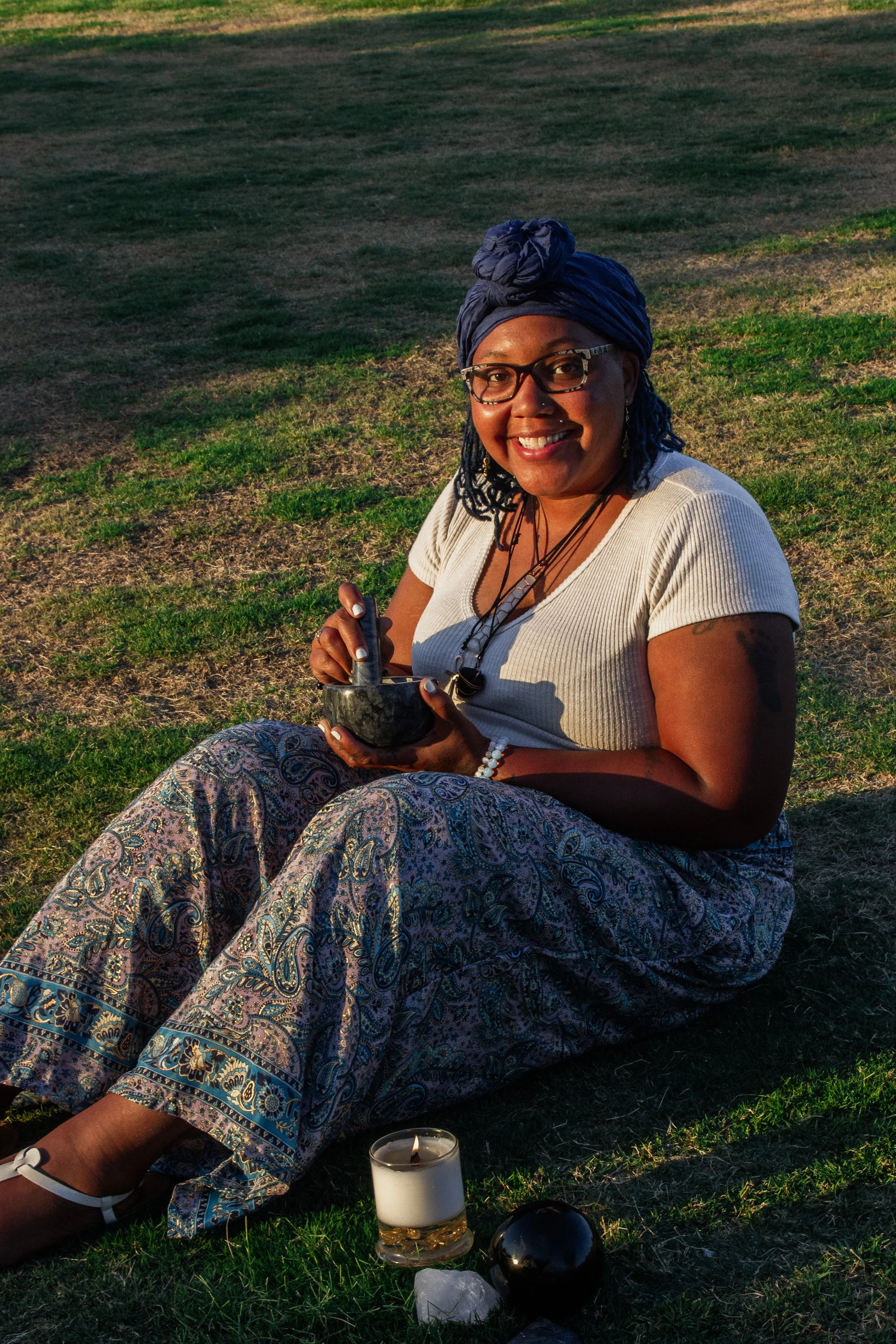 Mya from Cyan Candle Co. sitting on grass outdoors, smiling, wearing glasses, a headwrap, patterned pants, a white top, and multiple necklaces. She holds a small bowl and a pestle. Nearby on the grass are a candle, a black sphere, and some crystals.