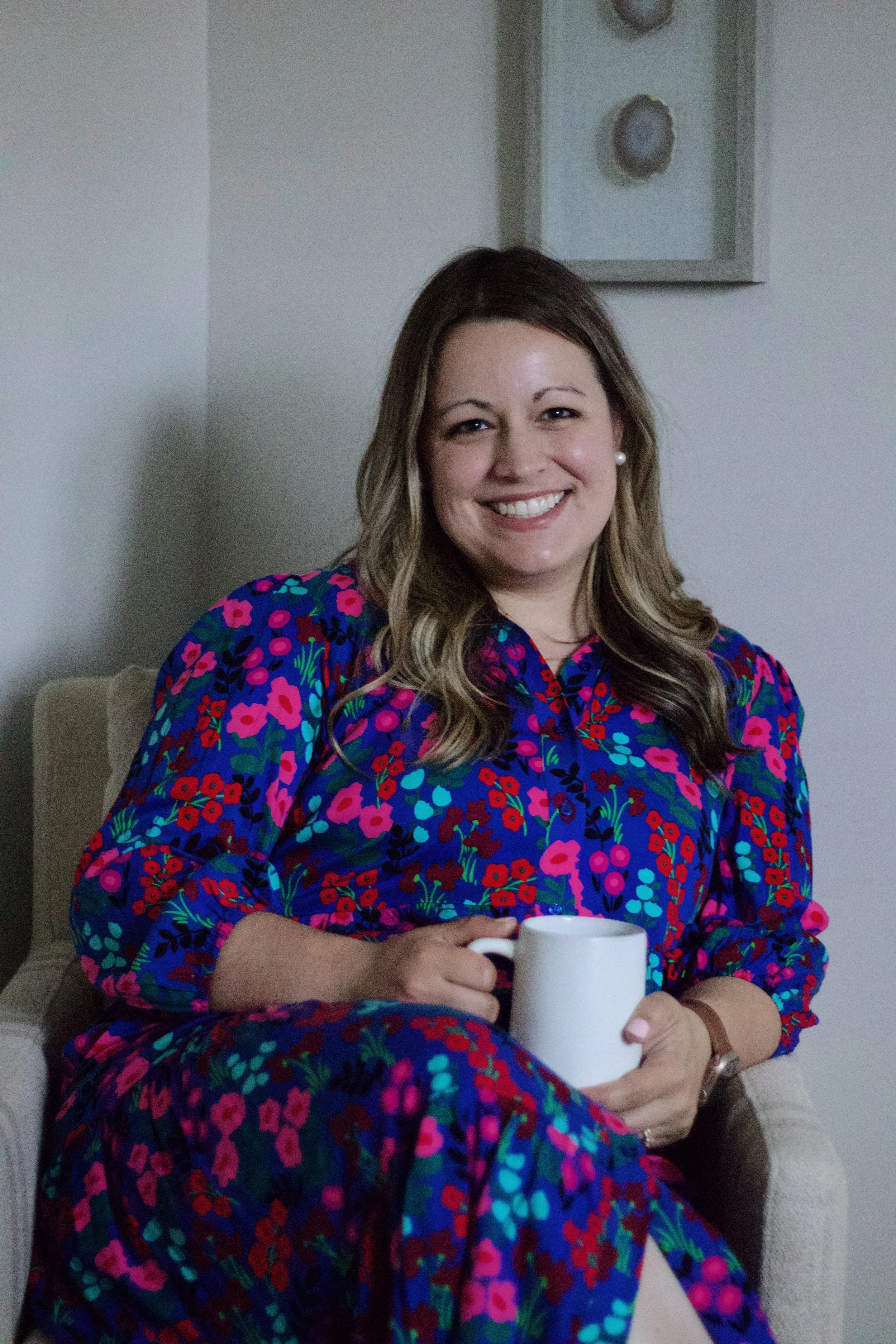 A woman with shoulder-length light brown hair, wearing a colorful floral dress, sitting in a beige armchair, holding a white mug, smiling at the camera.