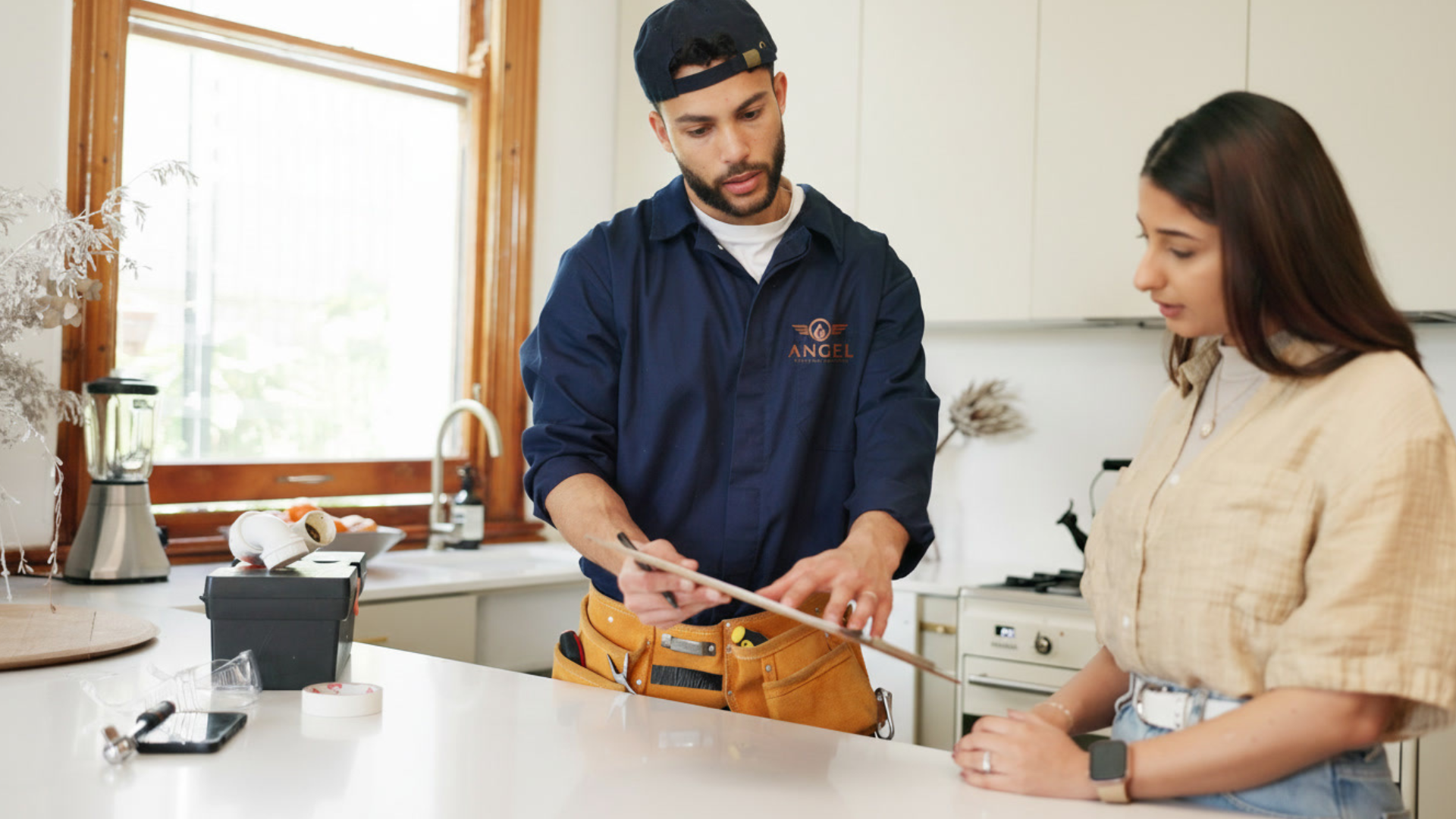 A professional plumber from Angel Sewer and Plumbing wearing a uniform and tool belt, showing a clipboard to a customer in a bright kitchen.