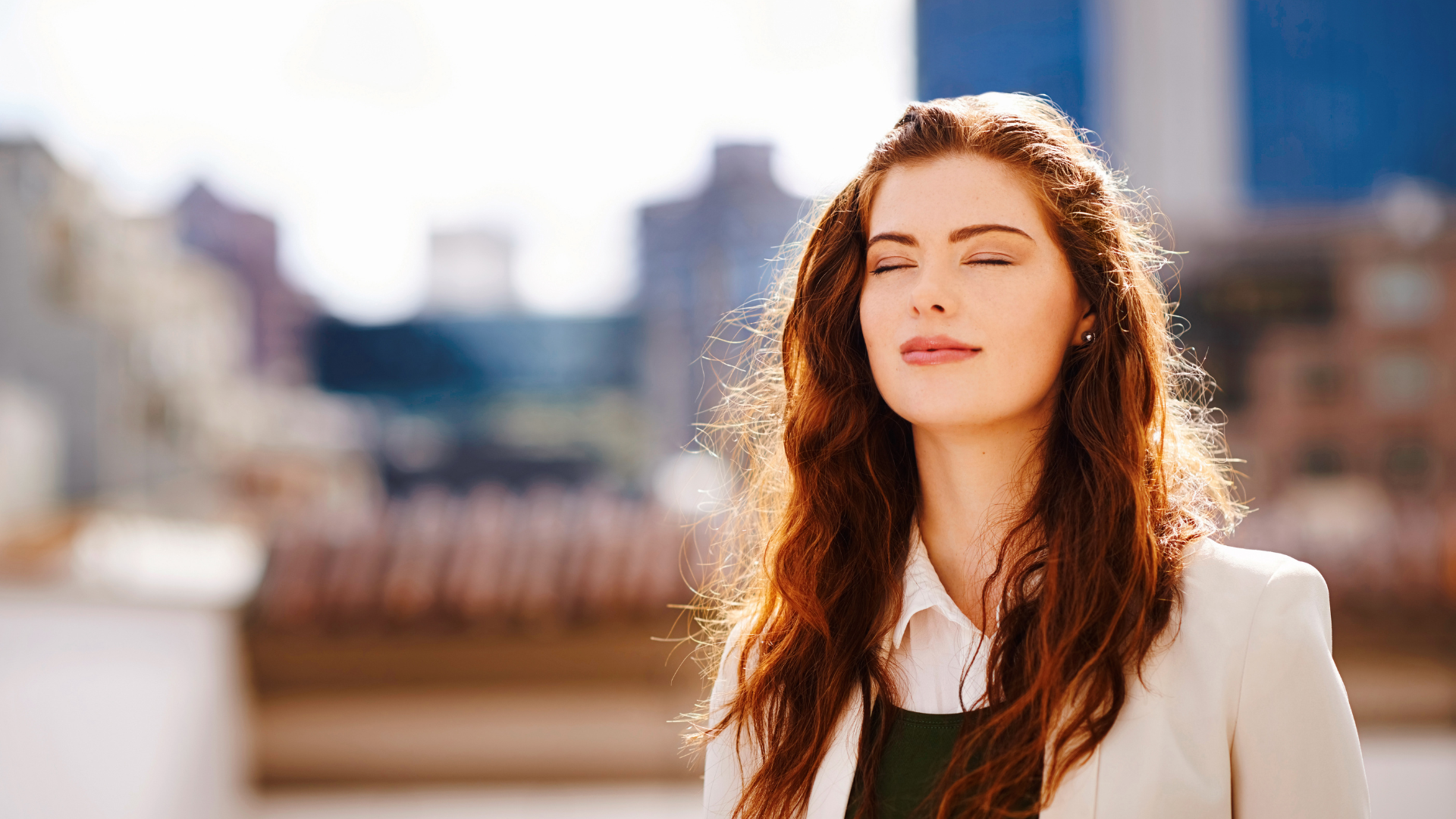A young woman with long, wavy red hair and closed eyes standing outdoors on a bright day with a blurred cityscape background.