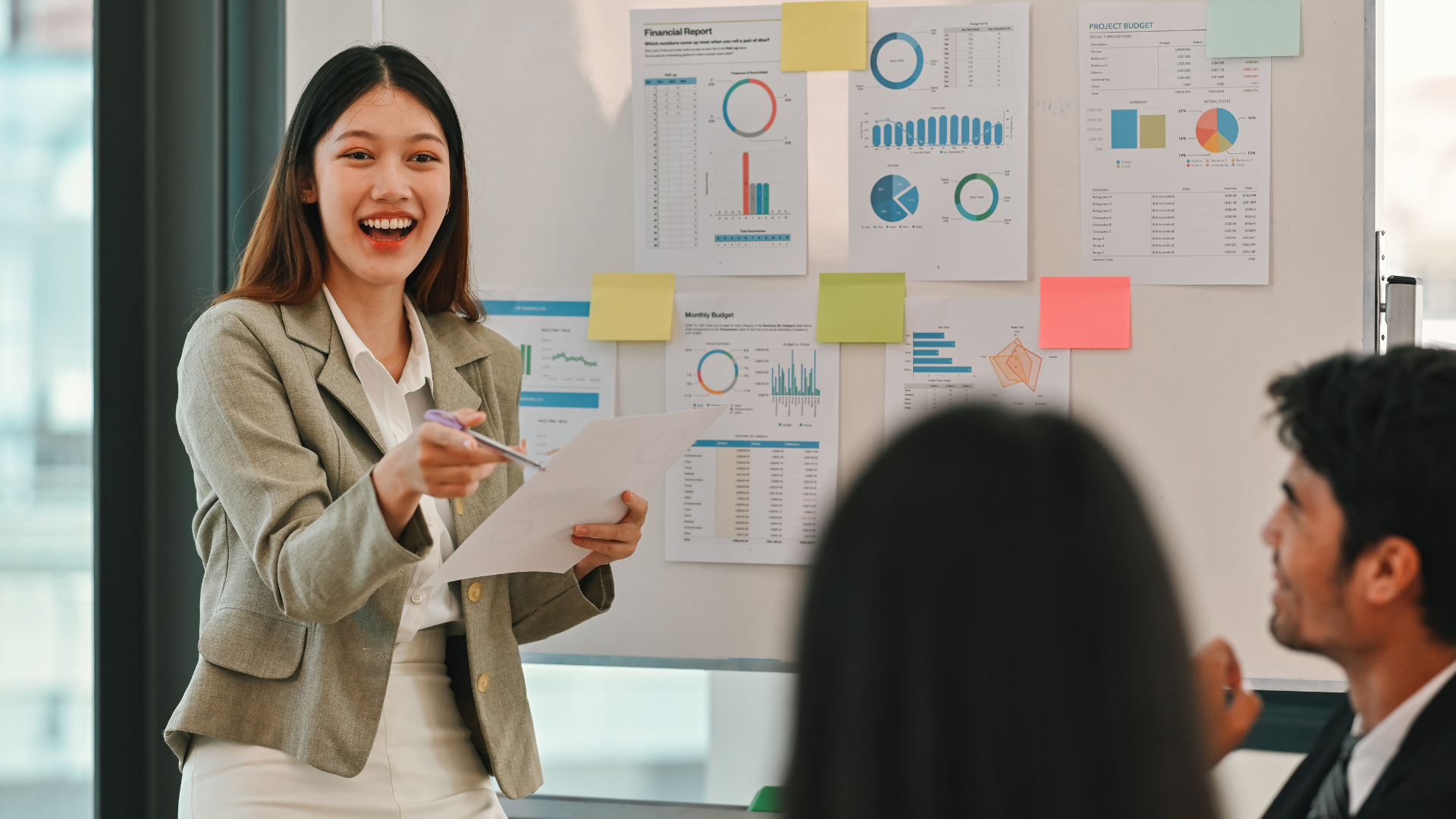 A woman in a beige blazer and white shirt holding a paper and pointing, smiling during a presentation in a meeting room with charts and graphs displayed on a wall.