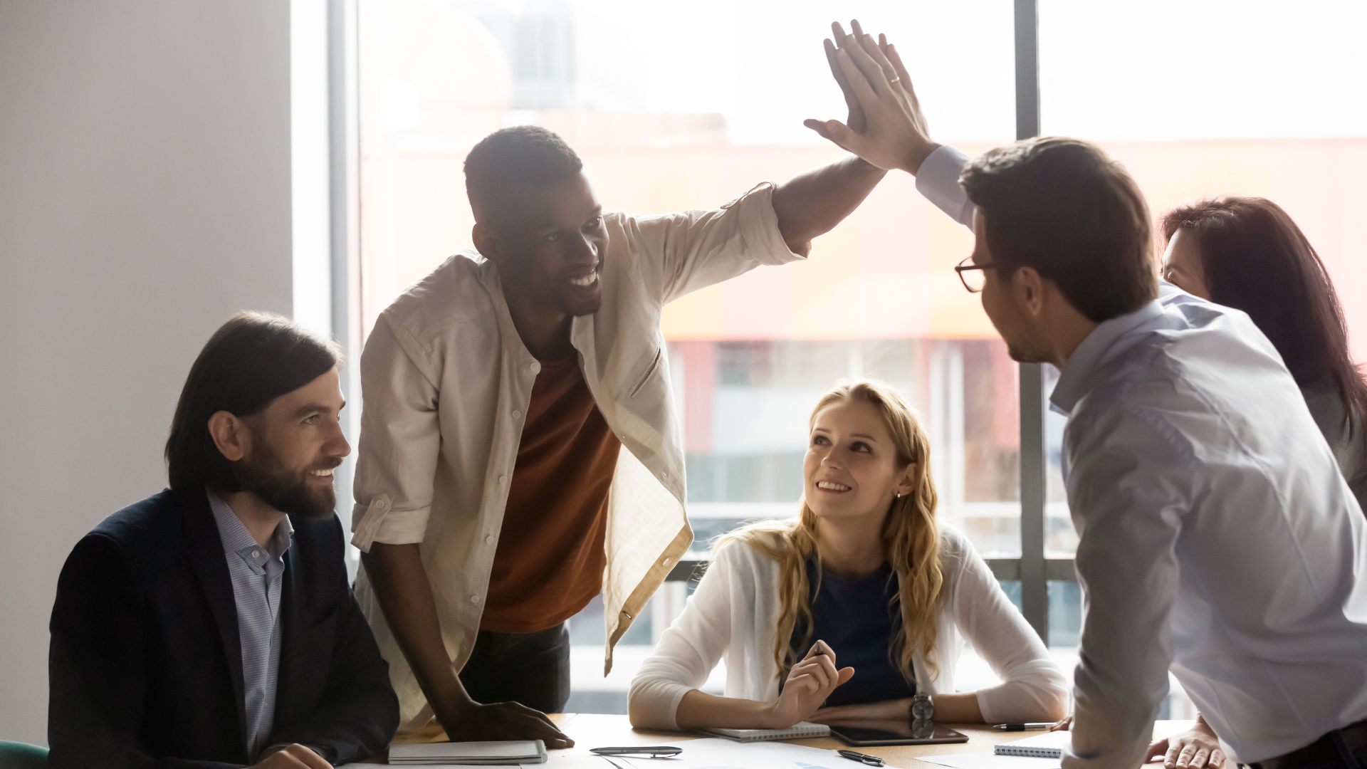 People in a business meeting giving a high five, smiling and engaging with each other in a conference room with large windows.