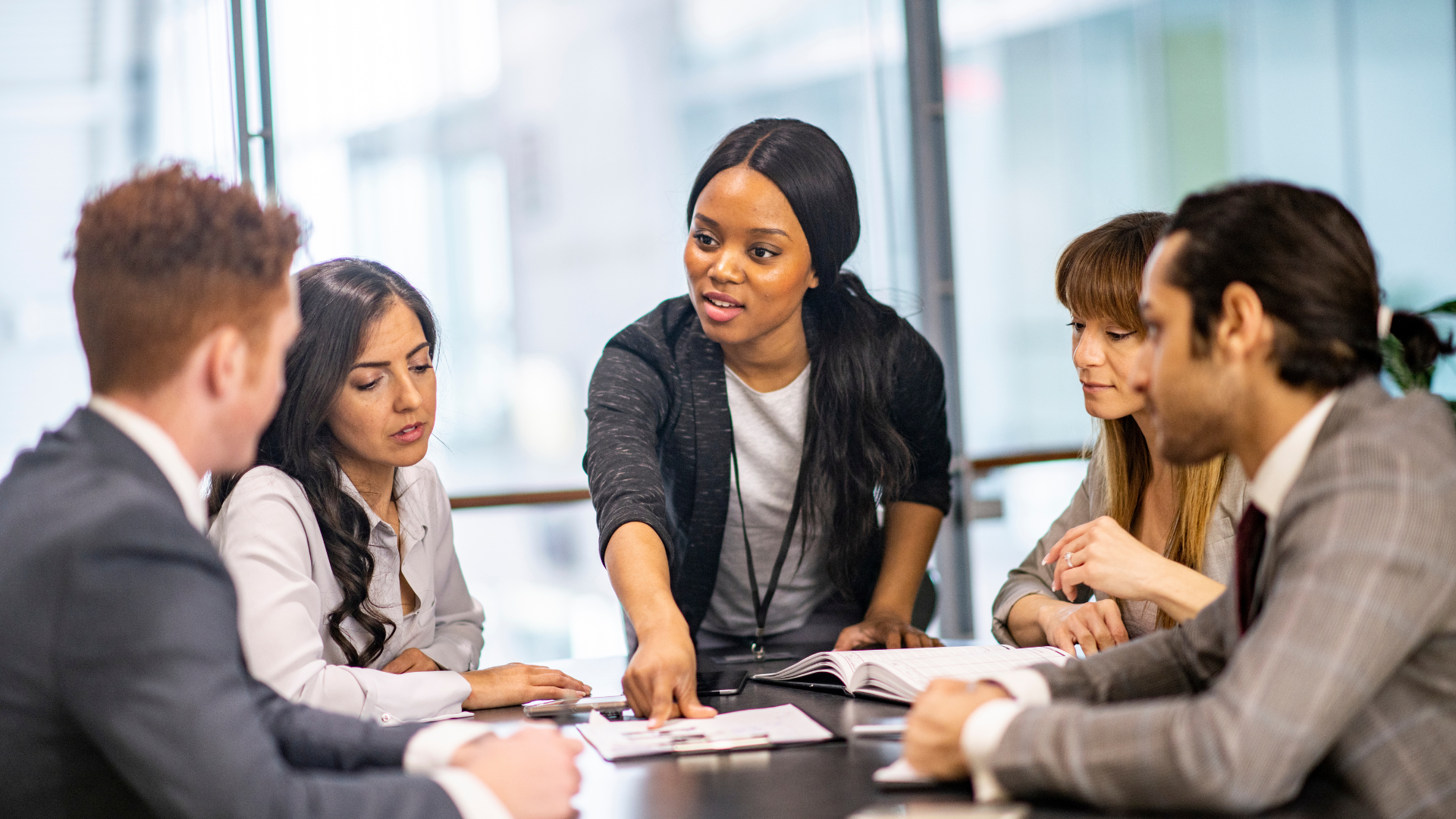 A diverse group of five professionals in a meeting, with a woman standing and pointing at documents on the table, discussing in an office setting.