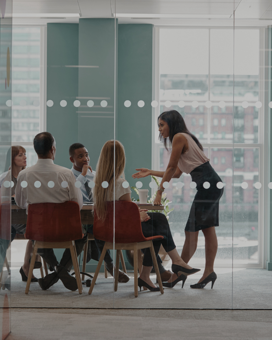 Businesswoman standing and talking to a seated group in a modern conference room with large windows.