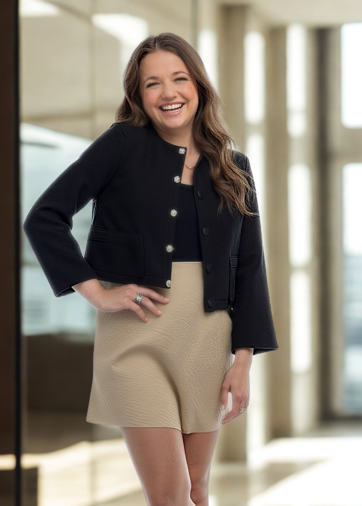 A woman with long brown hair smiling and posing indoors, wearing a black jacket and a beige skirt.