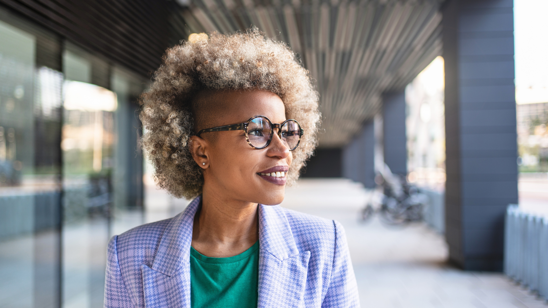 An older woman with curly gray hair, glasses, and earrings, smiling and looking to her left outdoors.