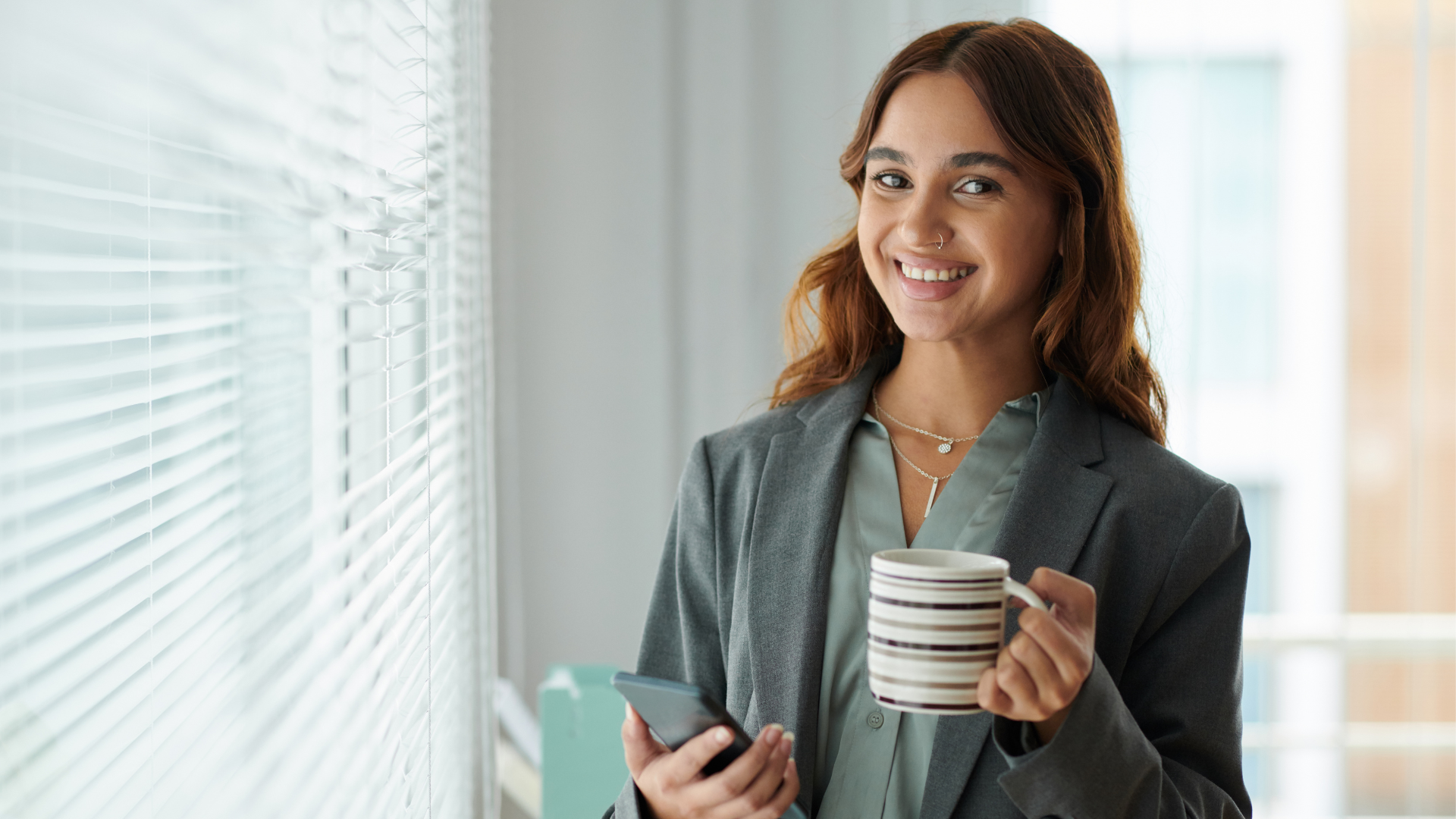 A smiling woman with reddish-brown hair, wearing a gray blazer and light-colored blouse, holding a smartphone in one hand and a striped coffee mug in the other, standing near window blinds in an indoor setting.
