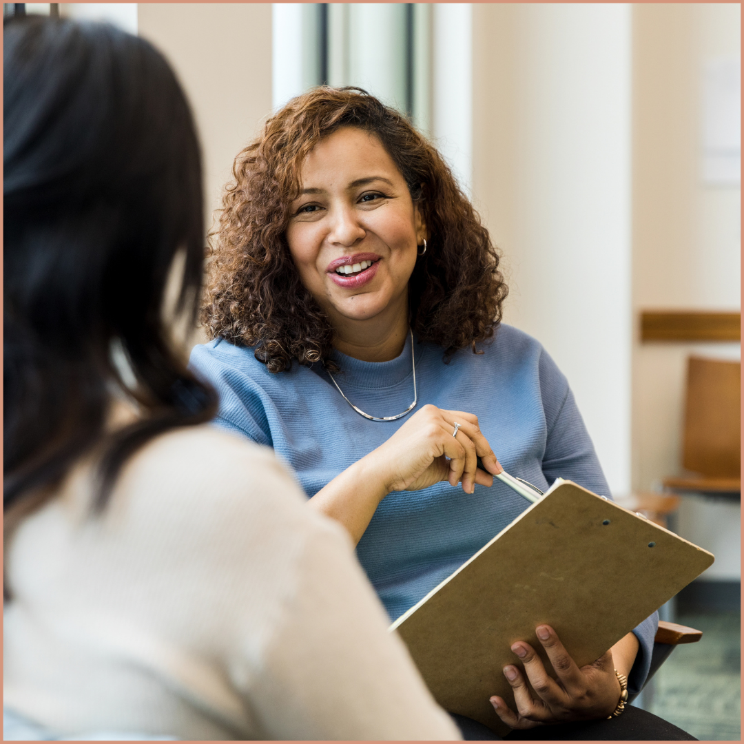 Two women sitting and talking in an indoor setting, with one woman smiling and holding a folder, engaged in a conversation.