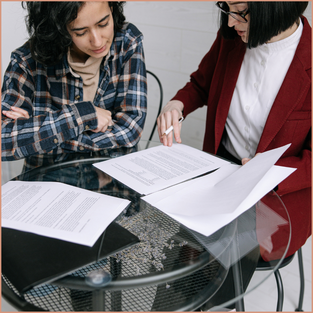 Two women sitting at a glass table reviewing documents, with one woman pointing at a paper.